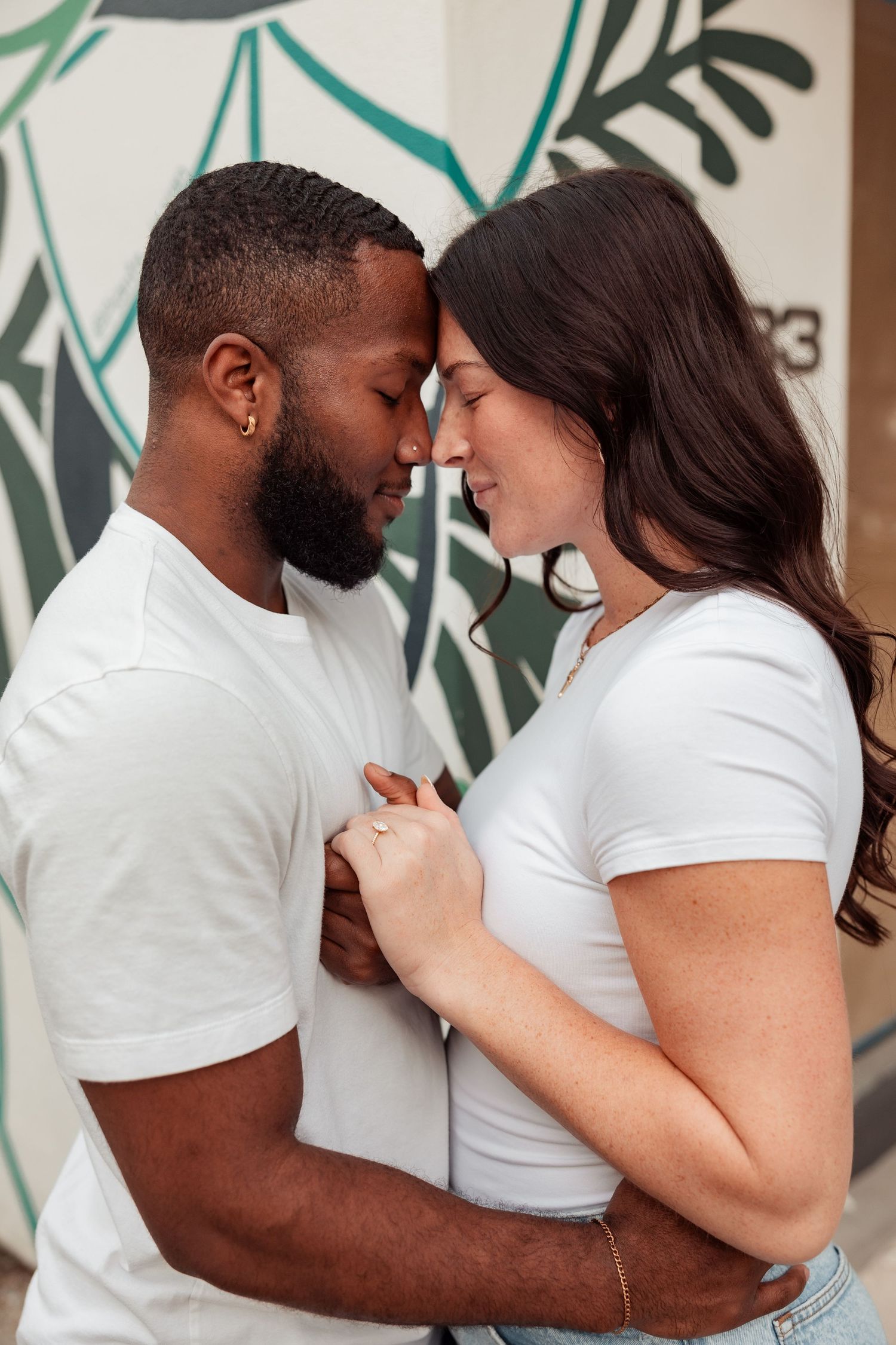 Romantic interracial couple touching foreheads tenderly in front of a tropical mural.