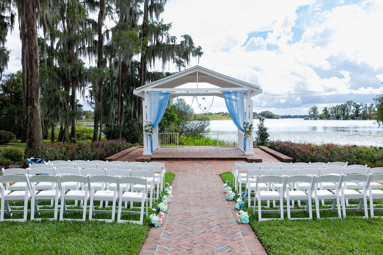 Elegant outdoor wedding ceremony setup with white chairs, brick aisle, and gazebo overlooking a serene lake.