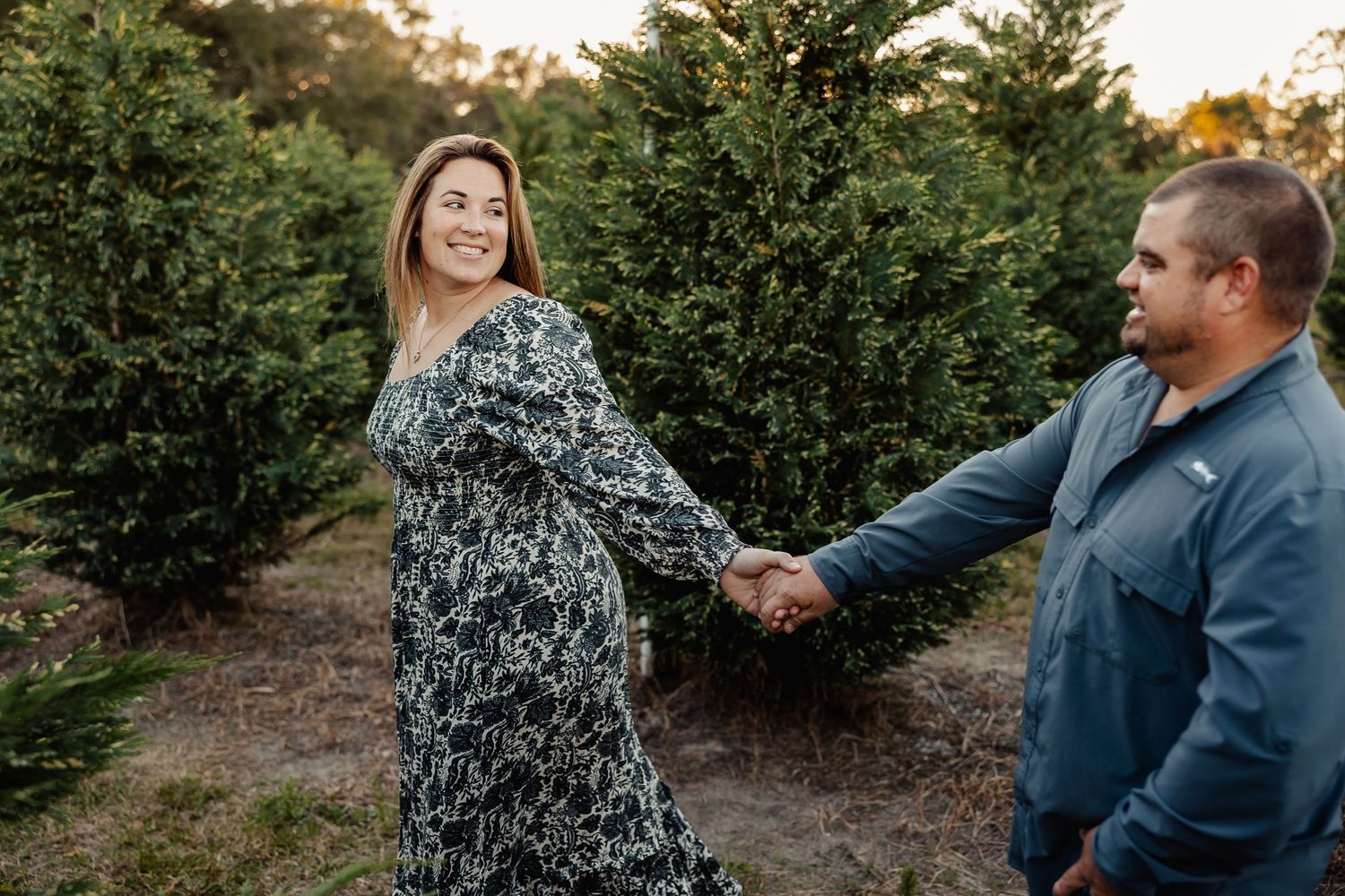 Woman in floral dress holding hands with man in blue shirt while walking through tree farm at sunset.