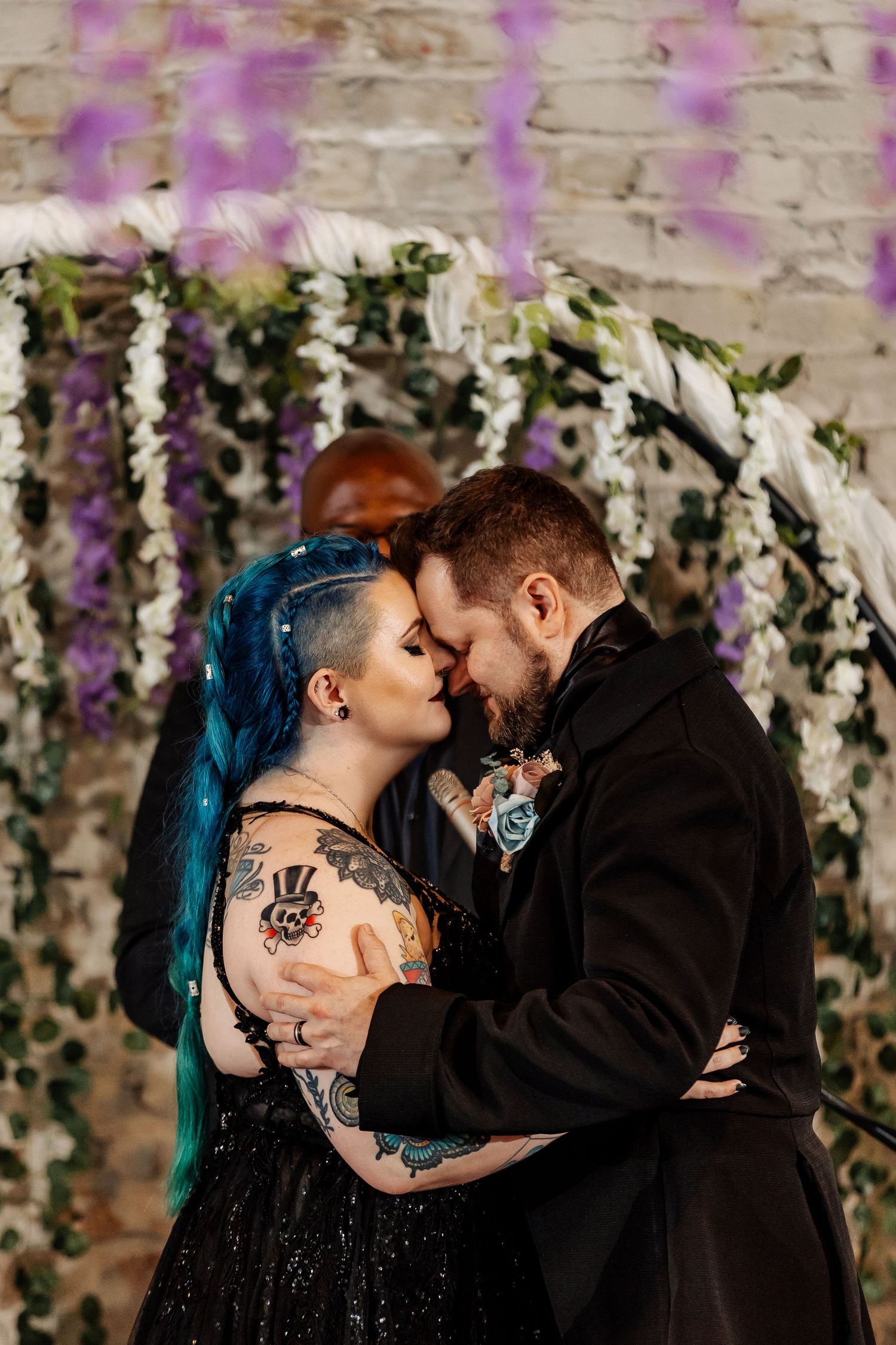 Tattooed bride with blue hair and groom share intimate moment before floral arch with wisteria.