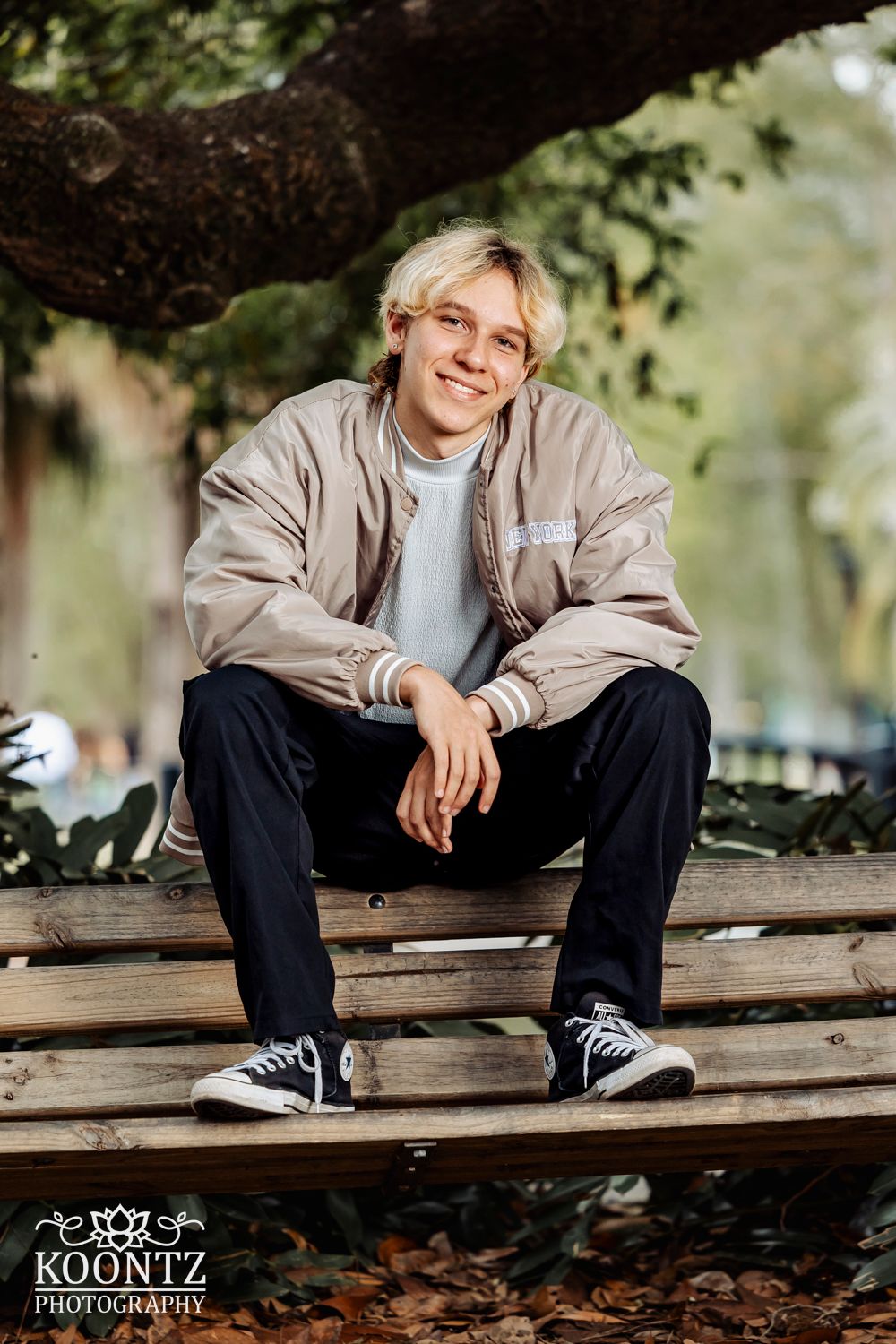 Professional outdoor senior photo shows casual pose sitting on wooden bench wearing beige jacket and Converse sneakers.
