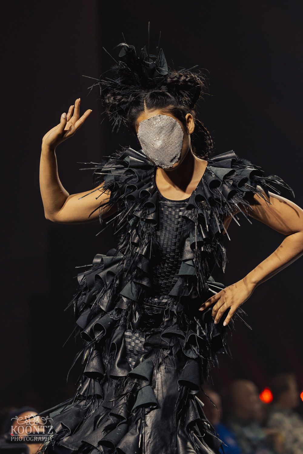Dramatic black feathered dress displayed on runway with theatrical lighting and dark backdrop.