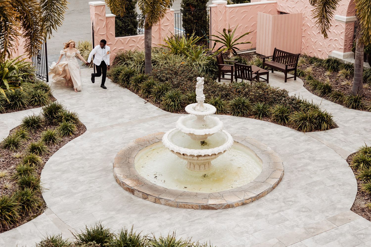 A white garden fountain with running water surrounded by manicured landscaping and palm trees.