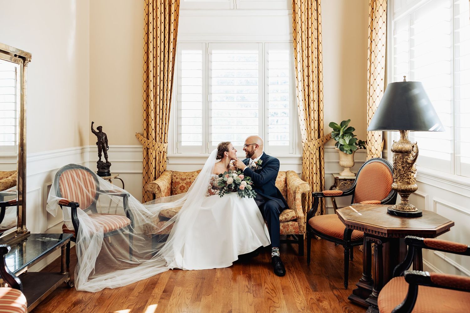 A couple in wedding attire sits together in an elegant vintage room with gold curtains and antique furniture.