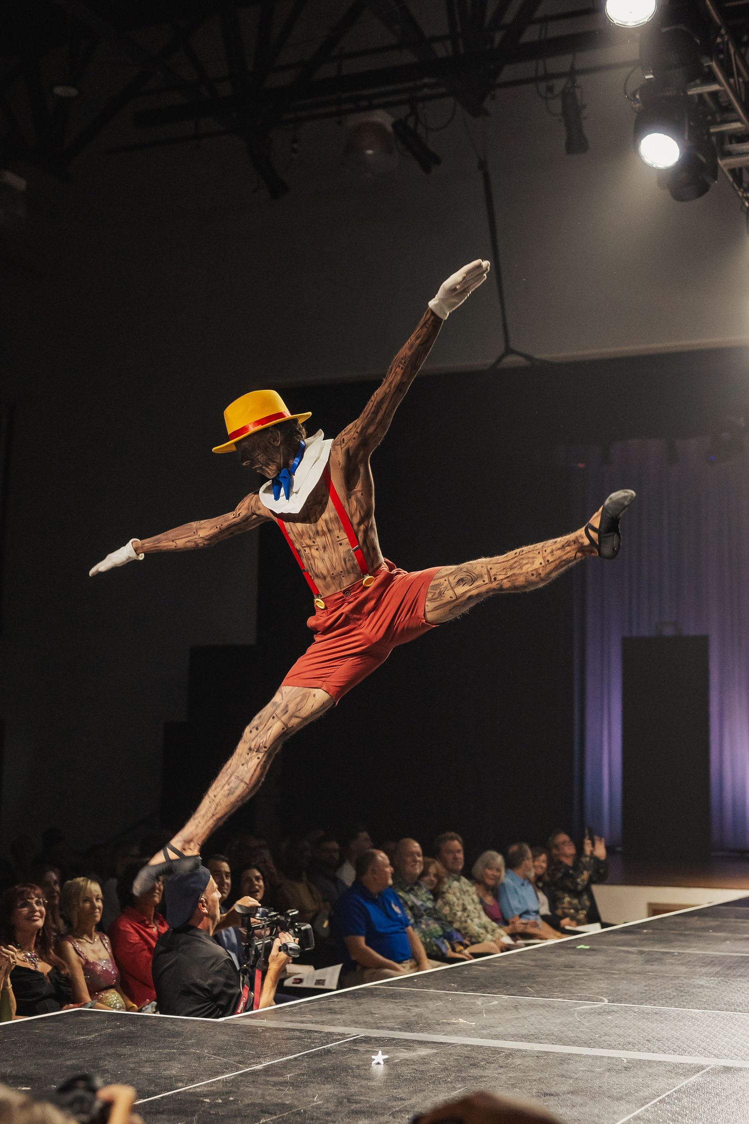 Tattooed performer in Pinocchio costume doing a split leap on a runway.