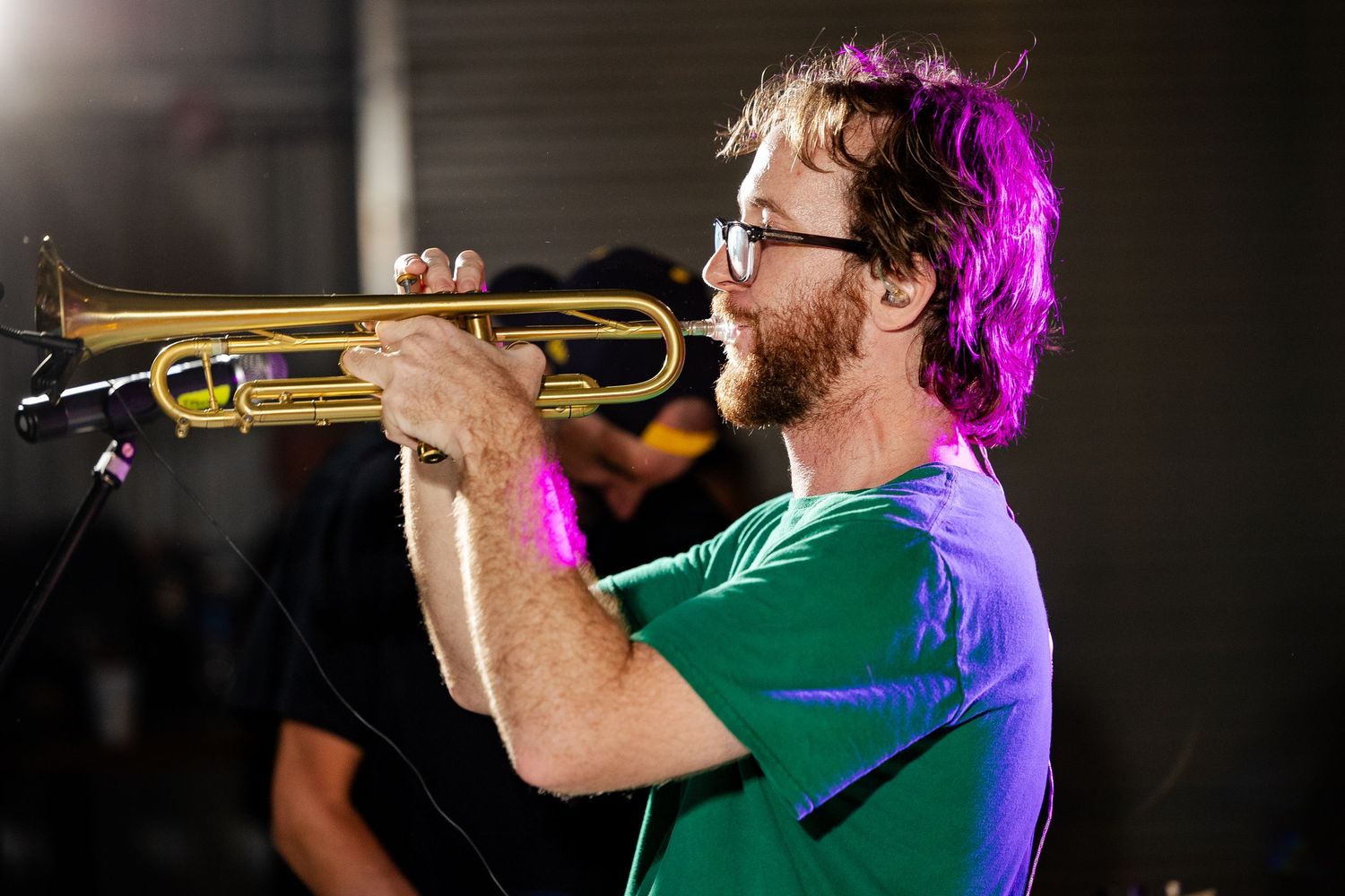 Male musician with beard playing trumpet on stage under purple stage lighting at live show.