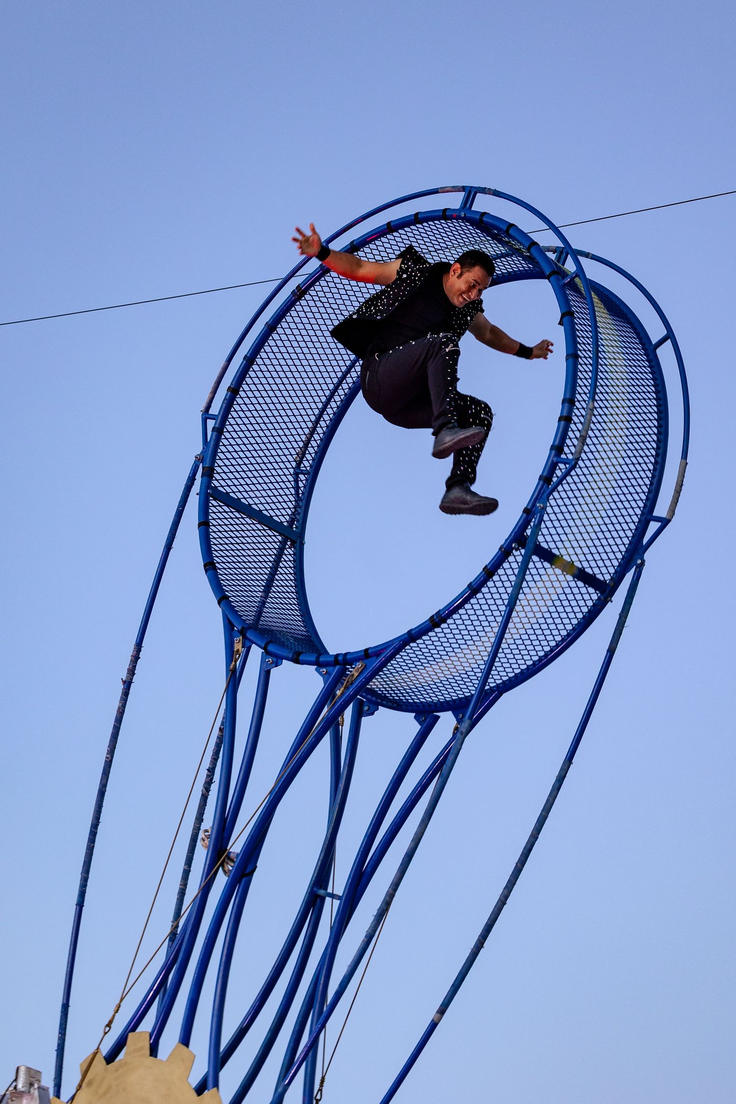 Daring circus performer running inside a giant blue wheel suspended high against a clear blue sky.
