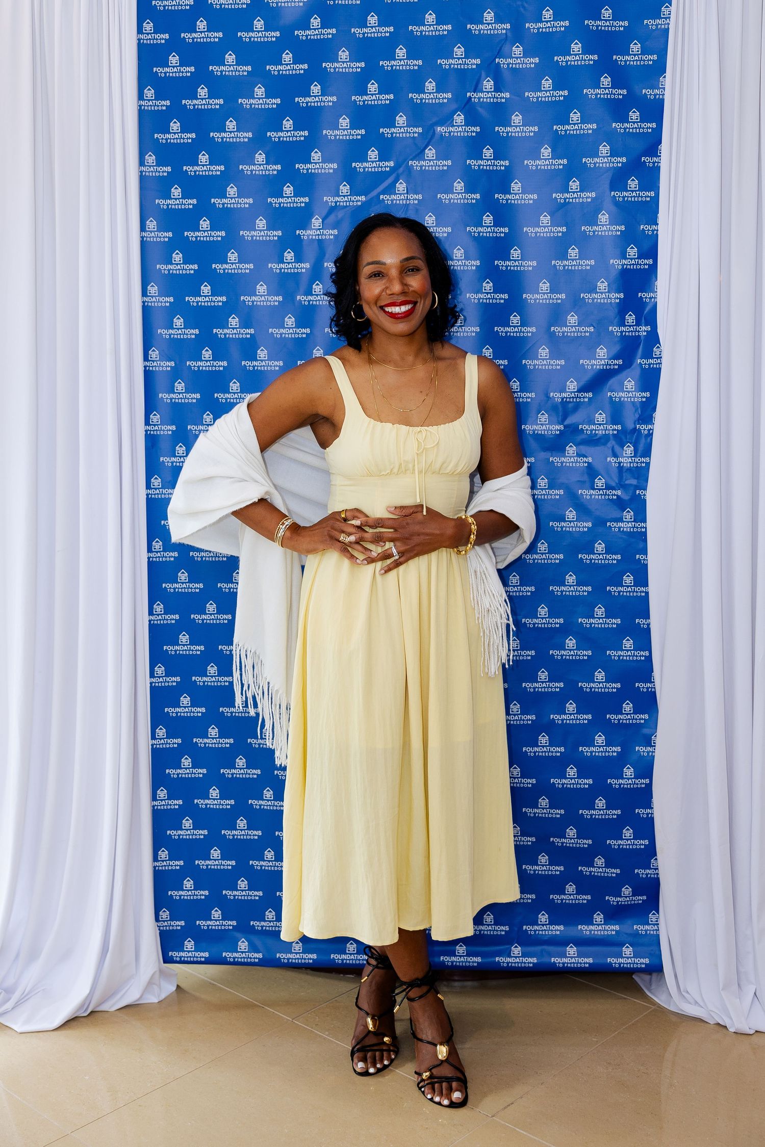 Smiling woman in yellow dress and white shawl poses in front of a blue branded step-and-repeat backdrop.