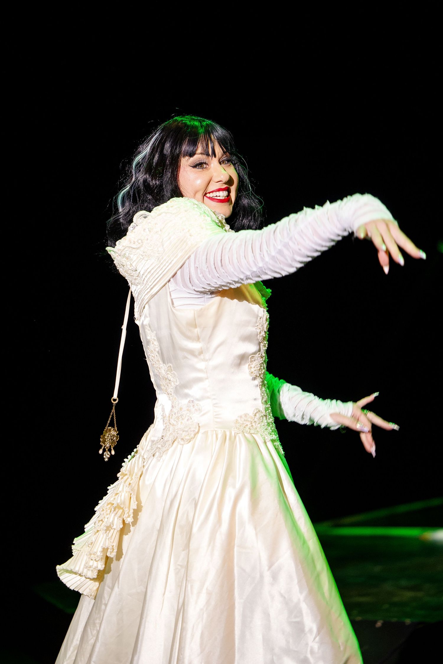 Burlesque performer in elegant white gown dancing on stage under green spotlight with arms extended.
