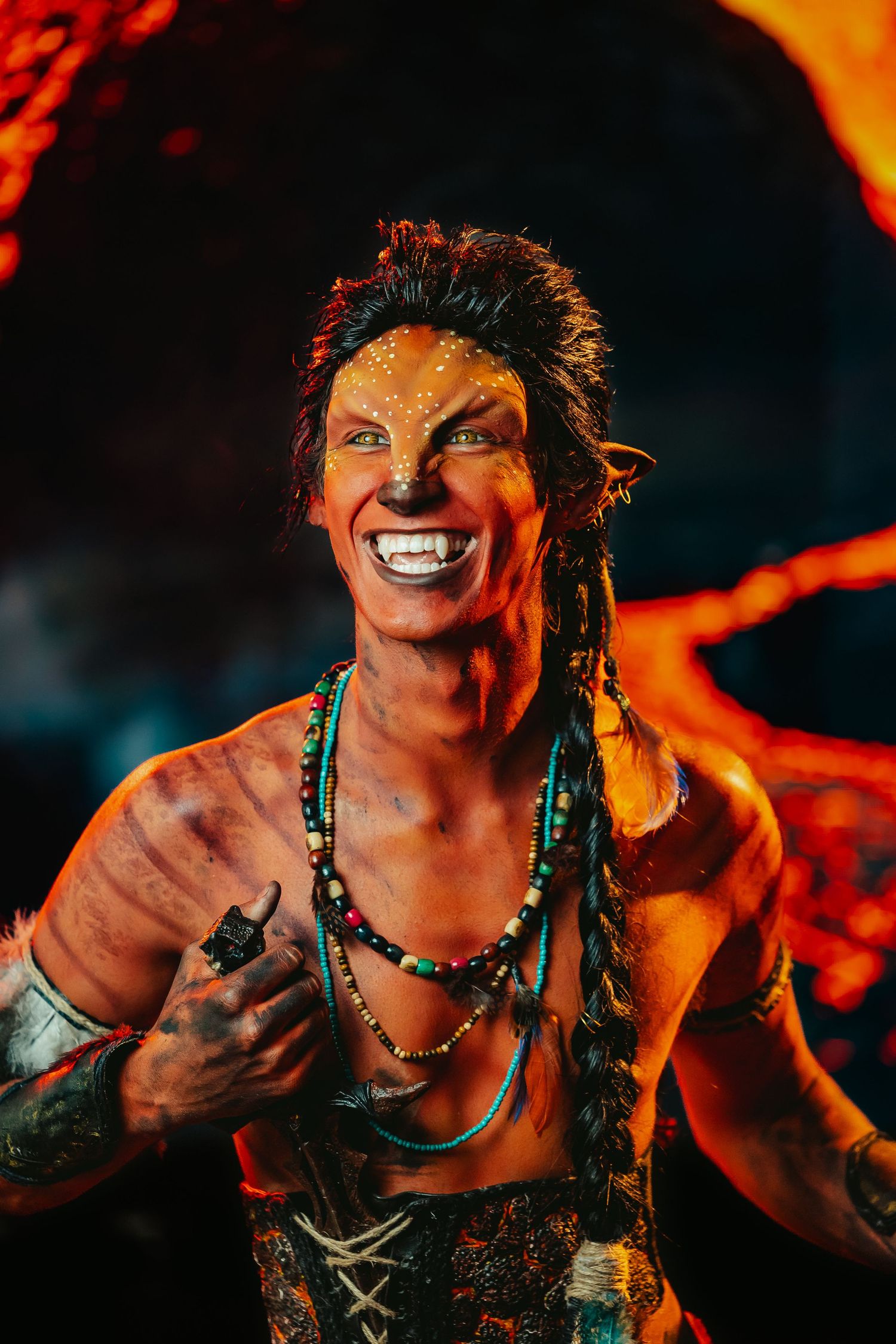 Smiling fantasy tribal character with spotted face paint, braids, and beaded necklaces against lava backdrop.