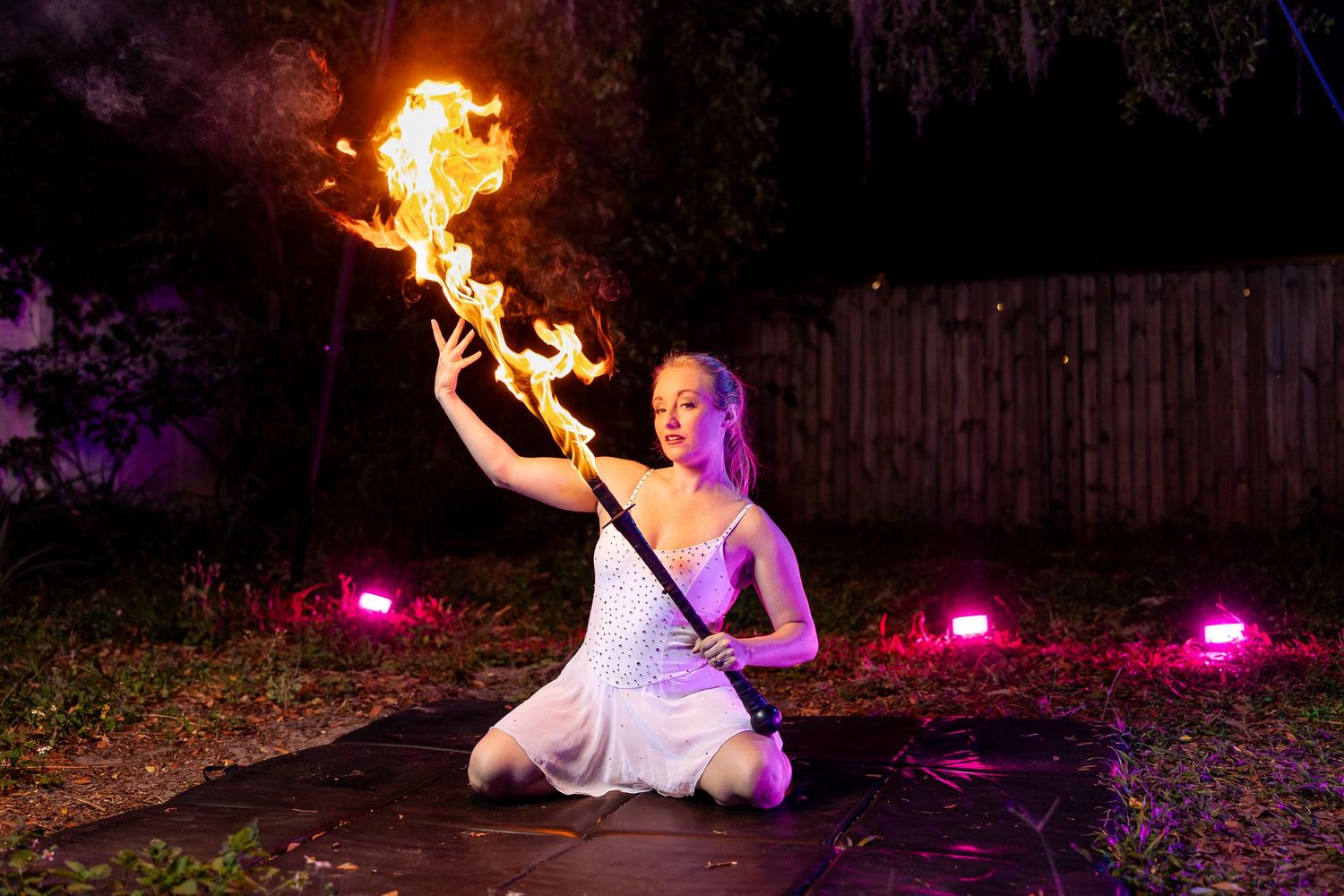 Blonde female performer kneeling on black mat holding a flaming fire staff aloft at night outdoors.