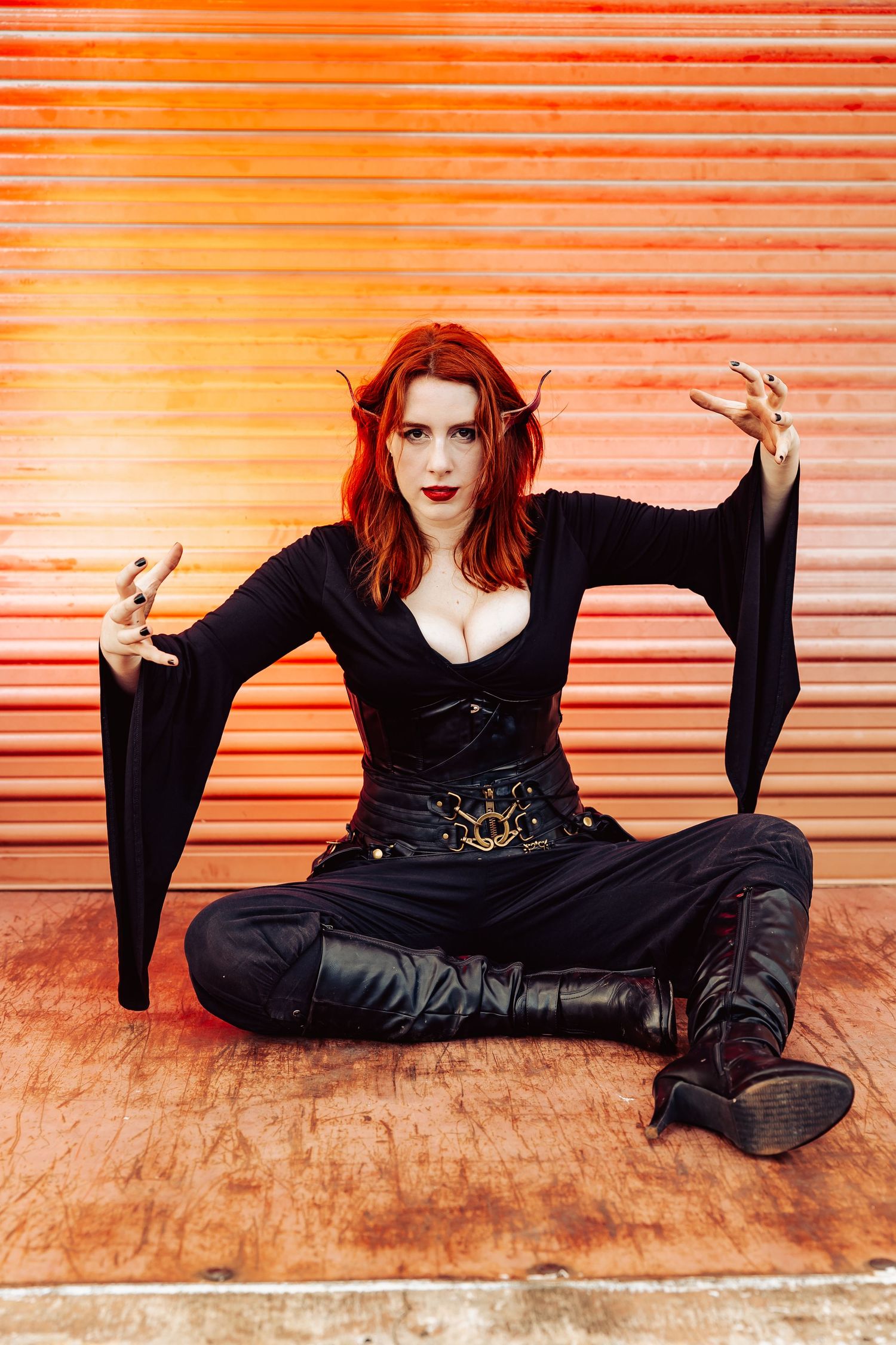 Redhead woman in dark elf cosplay with horns sits cross-legged posing dramatically before orange metal backdrop.