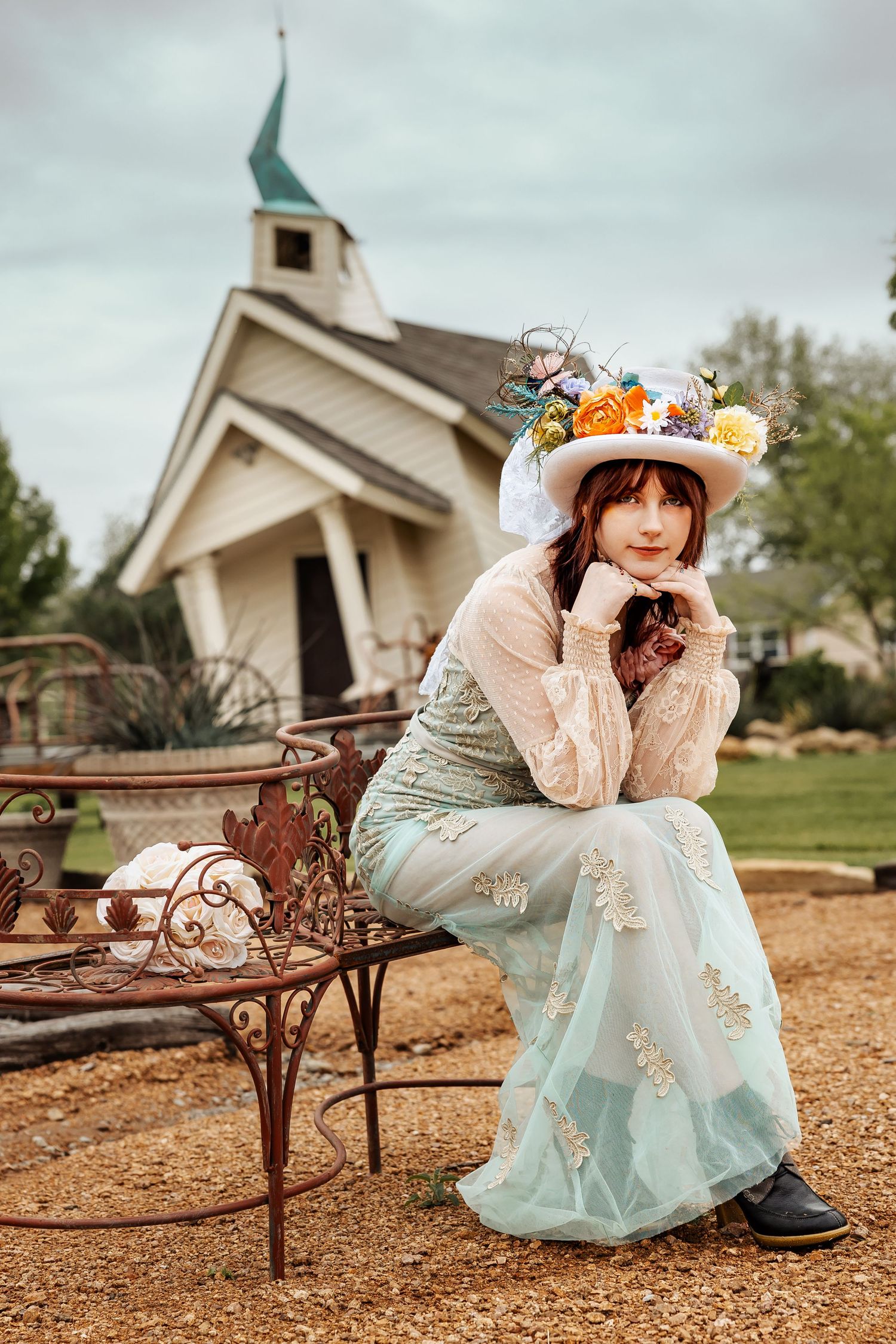 Woman in mint embroidered dress and floral hat sits on ornate iron bench before white chapel.