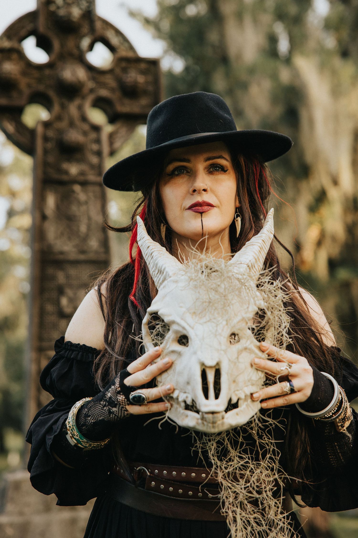 A woman in a black hat holds a horned animal skull in front of a stone cross in a cemetery.