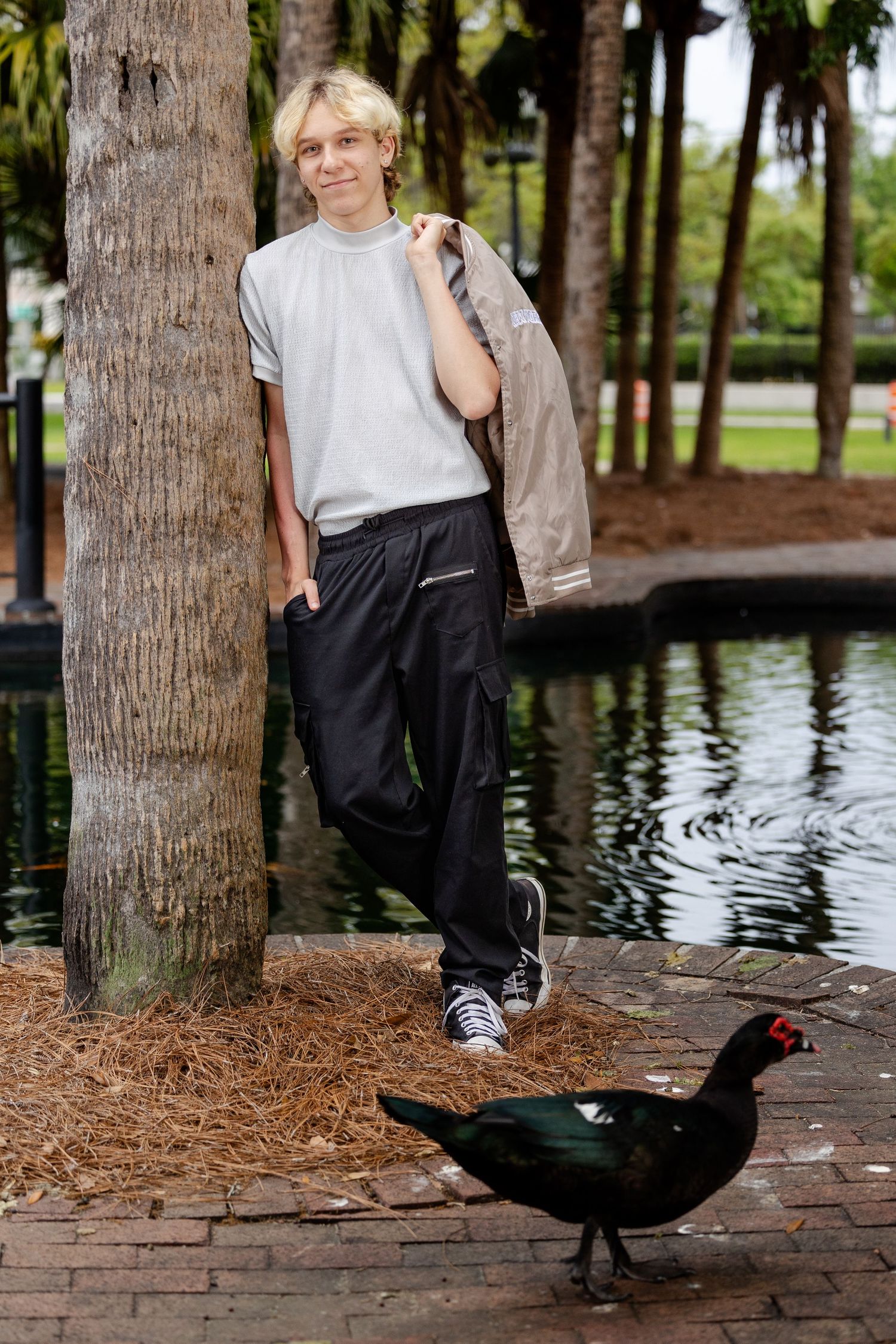 Teenage boy leaning against palm tree near pond in park, wearing cargo pants and white tee, with a black bird nearby.