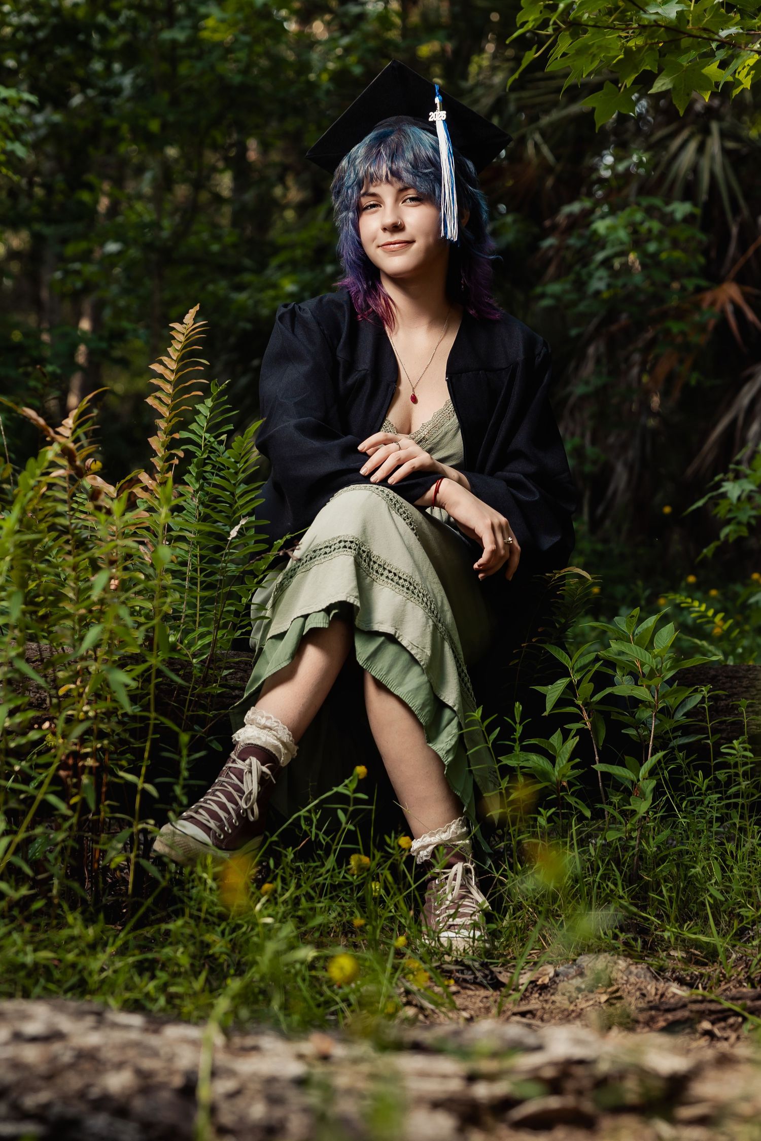 Young woman with blue-purple hair wearing graduation cap and gown sits among lush green ferns in a forest setting.