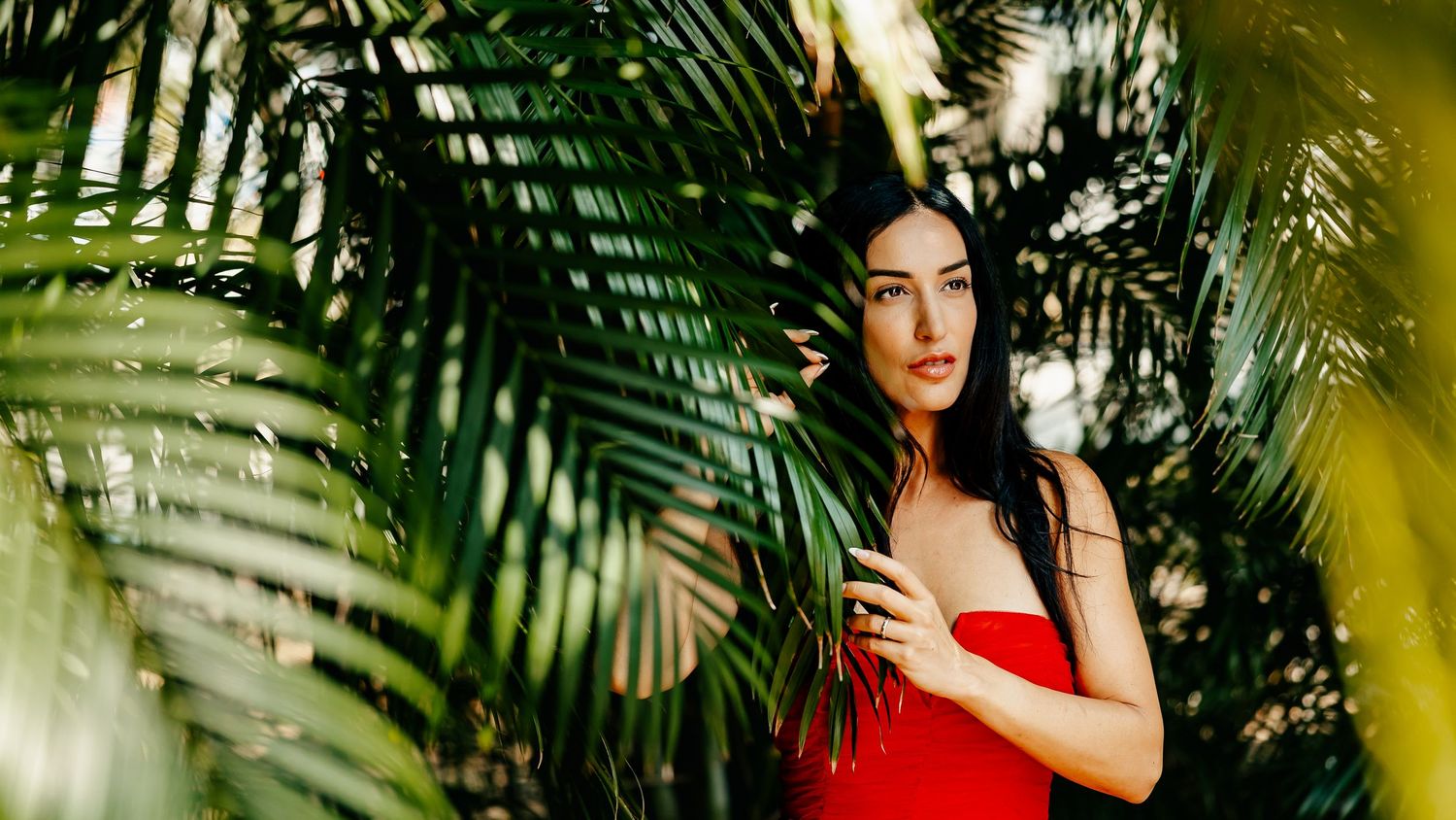 A woman in a red dress poses among lush tropical palm leaves in a vibrant outdoor setting.