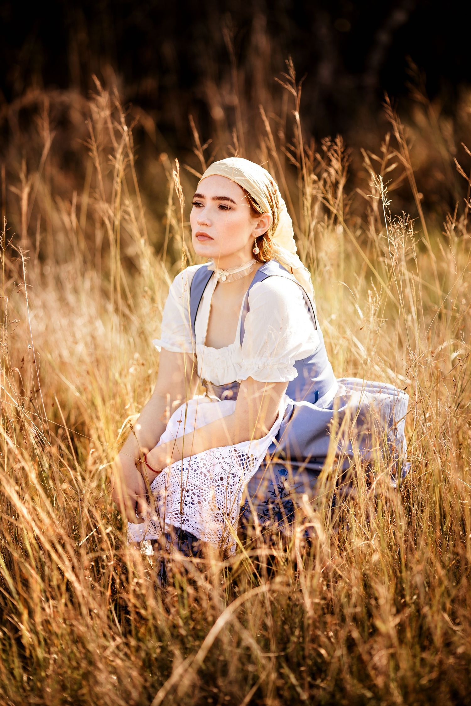 Young woman in vintage dirndl dress and headscarf sits gracefully among tall golden grass in warm sunlight.