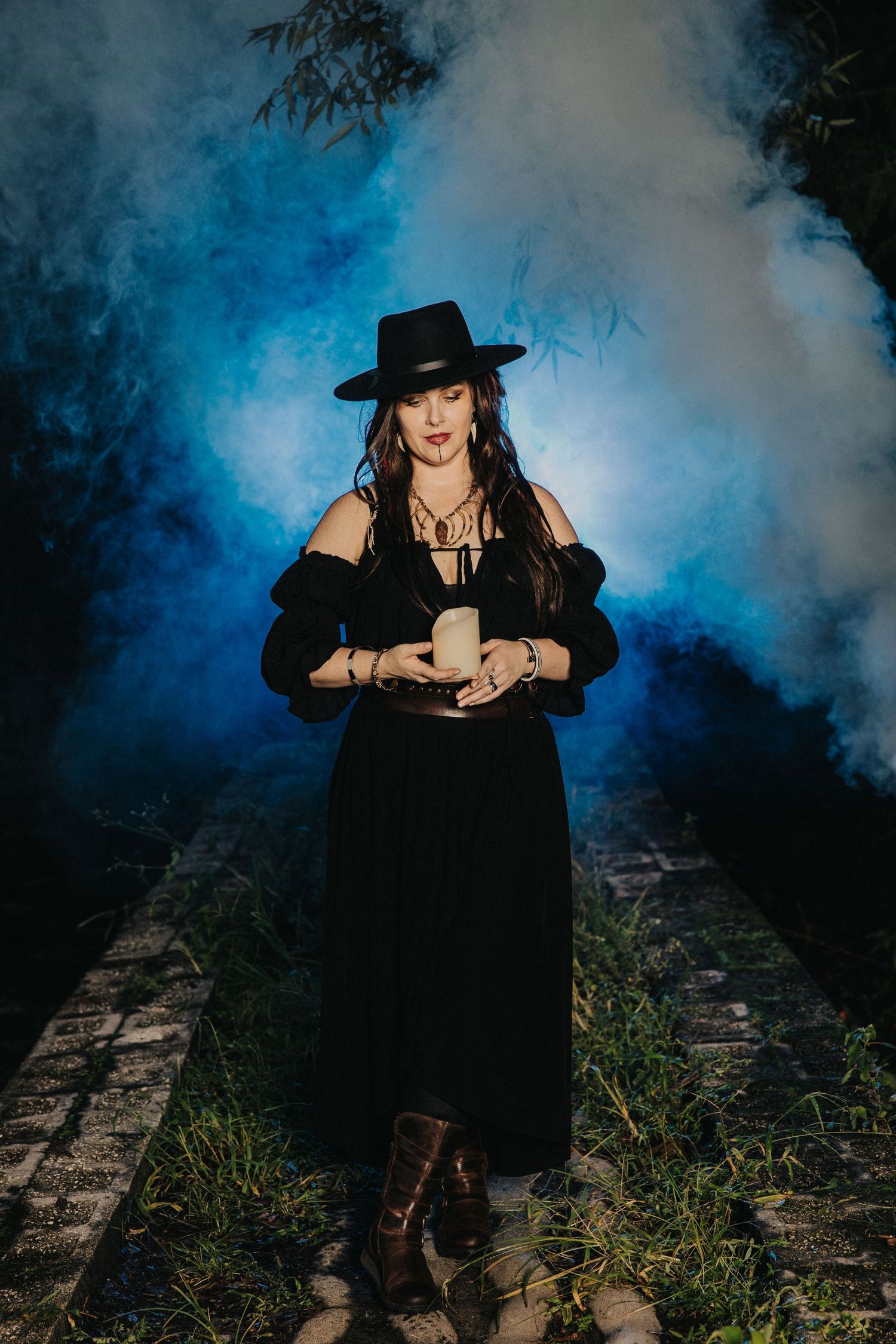 Woman in black off-shoulder dress and wide-brim hat holds a candle surrounded by blue smoke at night.
