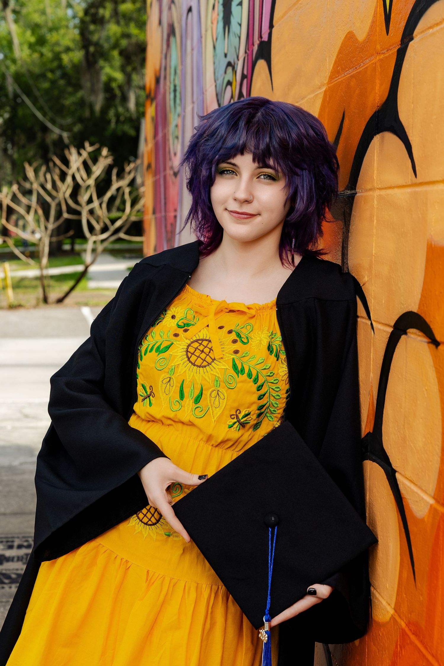 Young woman with purple hair in graduation gown and cap holds diploma, posing in yellow dress by colorful graffiti wall.