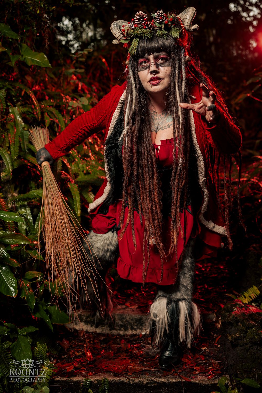 Woman in red dress with horns, dreads, and broom crouches in a dark forest in festive Krampus cosplay.