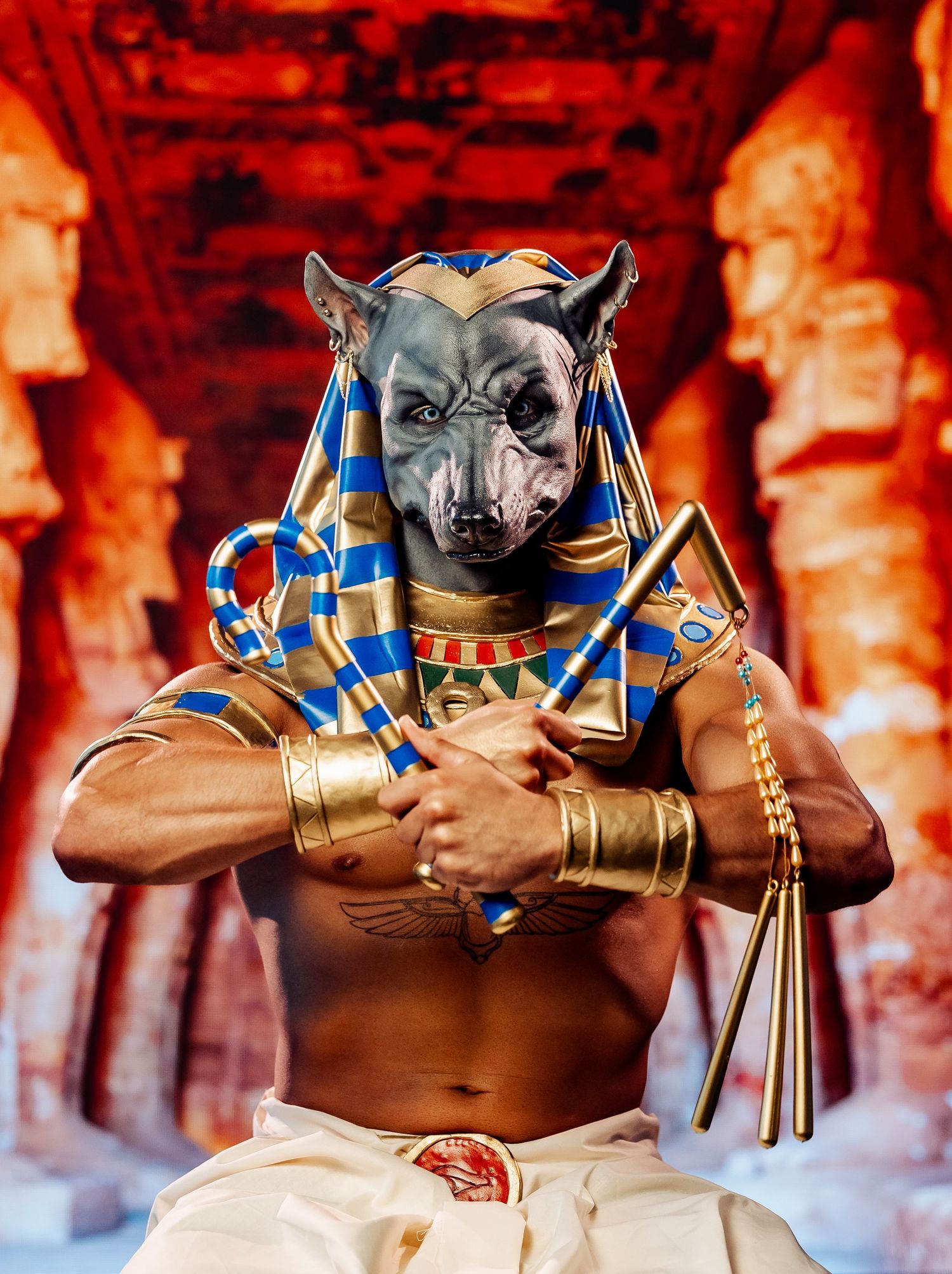 Costumed performer as Egyptian god Anubis with jackal mask holding golden crook in temple setting.