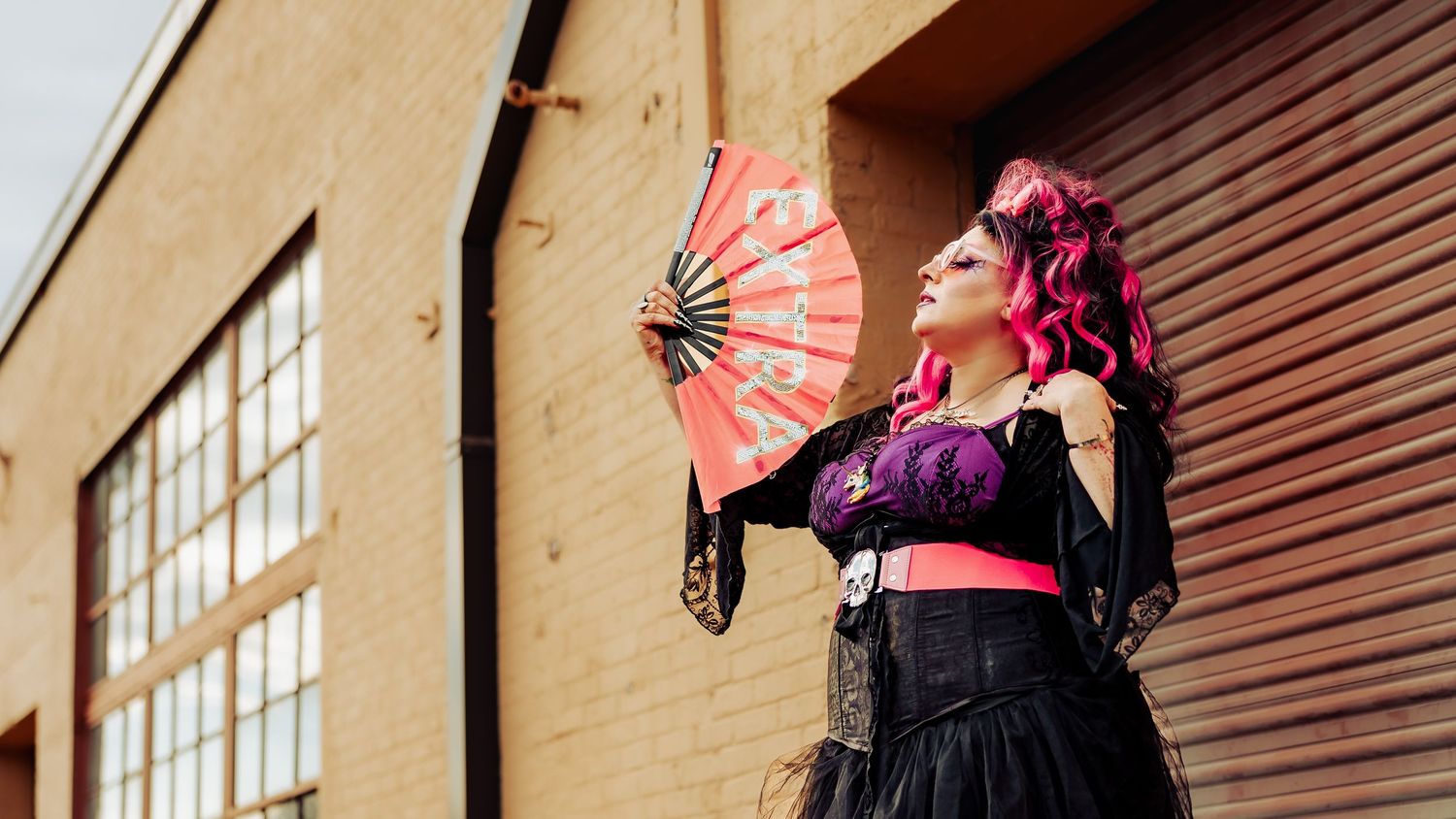 Woman in gothic black corset dress holds a red fan while posing outside a modern building.