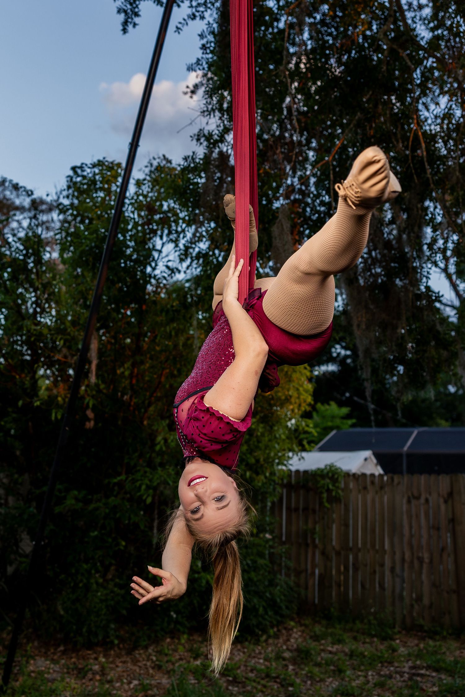 Smiling aerialist hangs upside down on red aerial silks among trees outdoors.