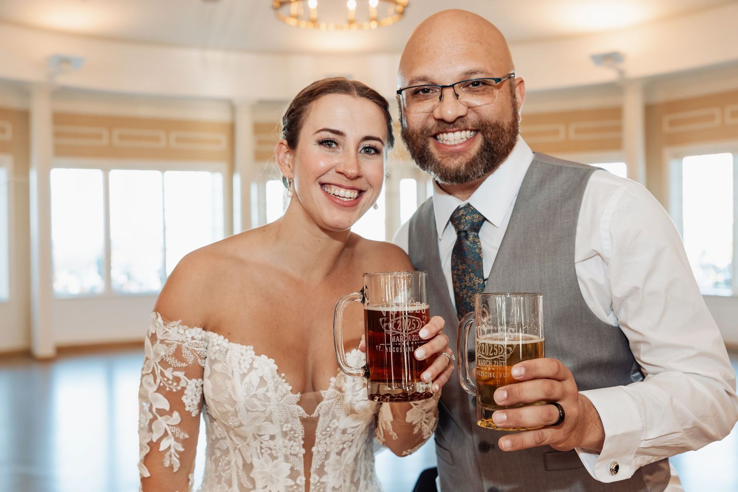 Smiling bride and groom toasting with beer mugs at their wedding reception indoors.