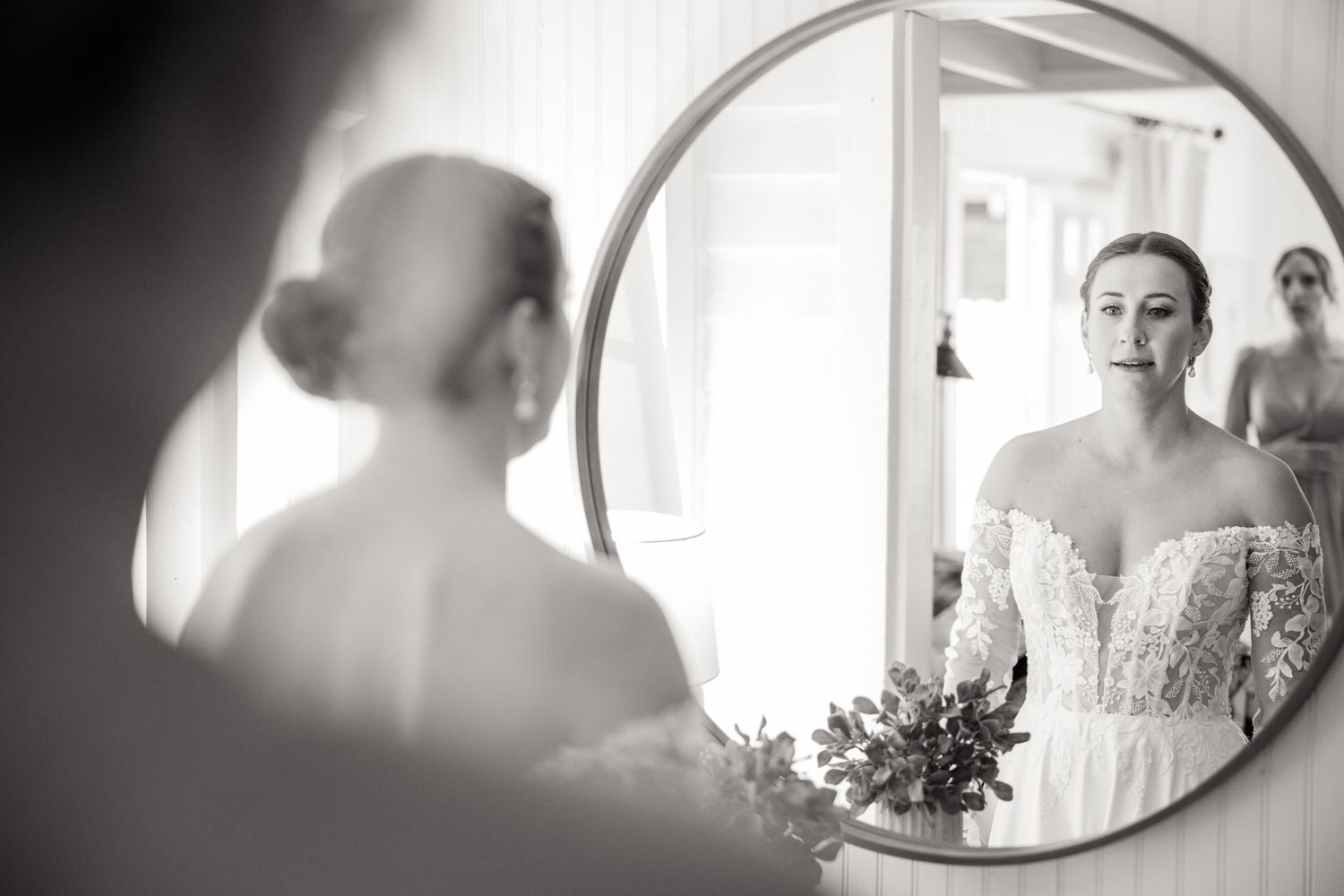 Black and white photo of bride in lace gown viewing her reflection in a round mirror.