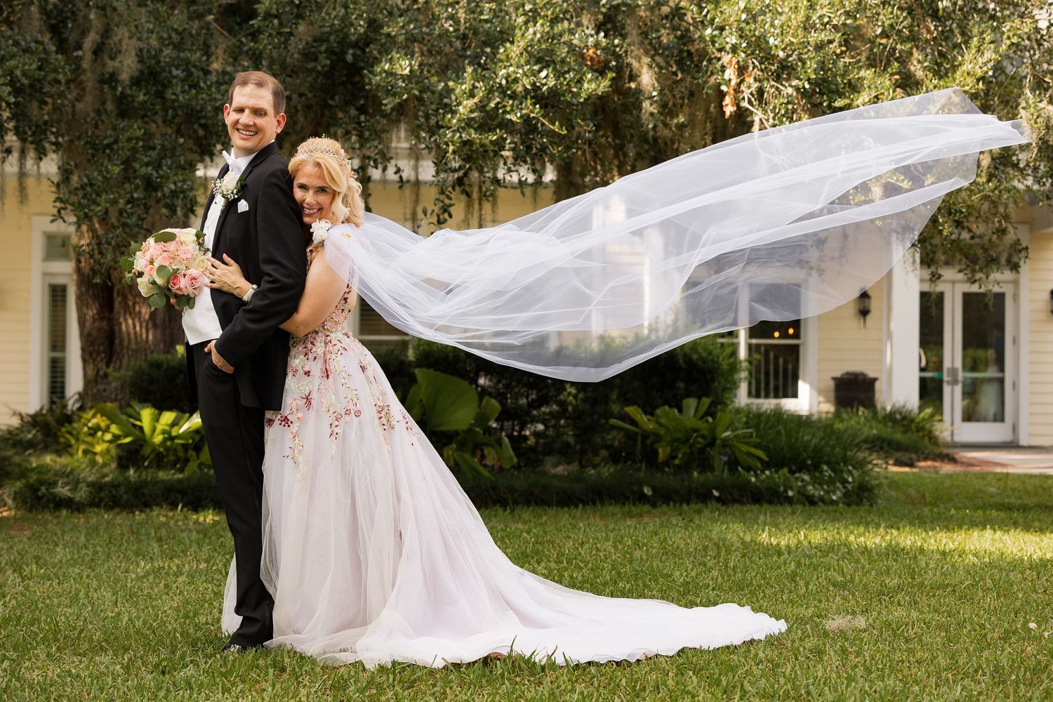 Bride and groom posing outdoors with flowing veil billowing in wind before elegant white estate.