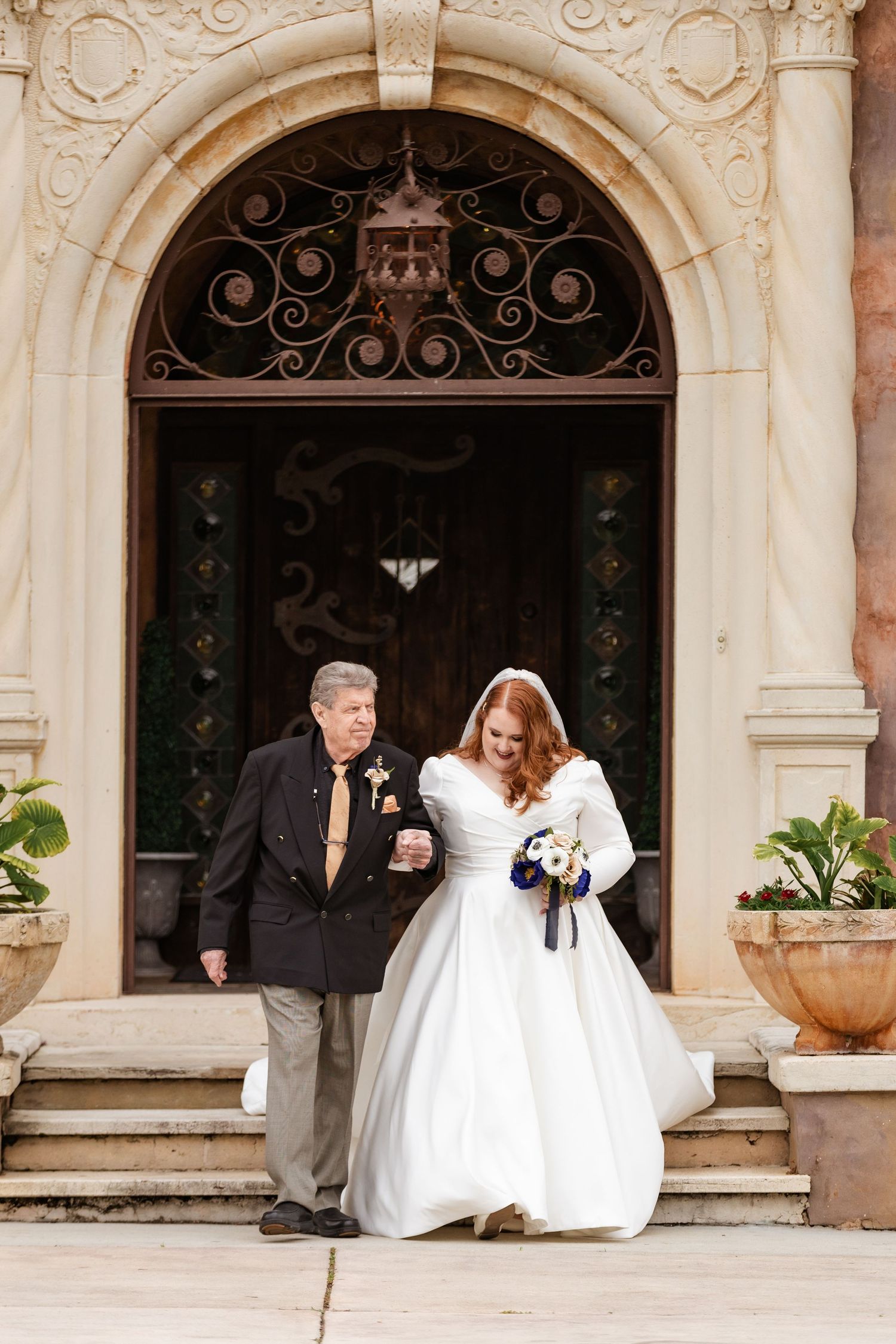 Father of the bride escorting his daughter in a white wedding gown down the steps of an elegant mansion.