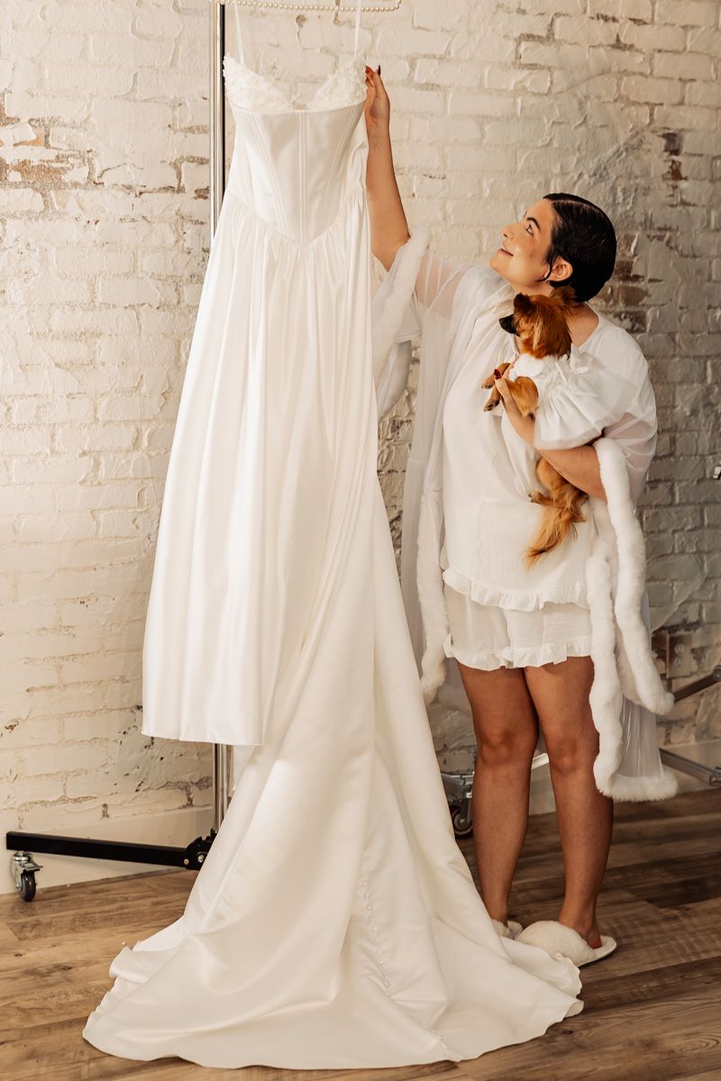 Bride in white robe holding a small dog while admiring her wedding gown hanging in a bright studio.