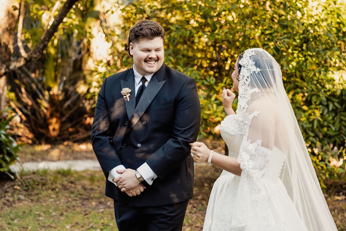 Groom in black suit smiles at bride in lace veil during outdoor wedding first look moment.