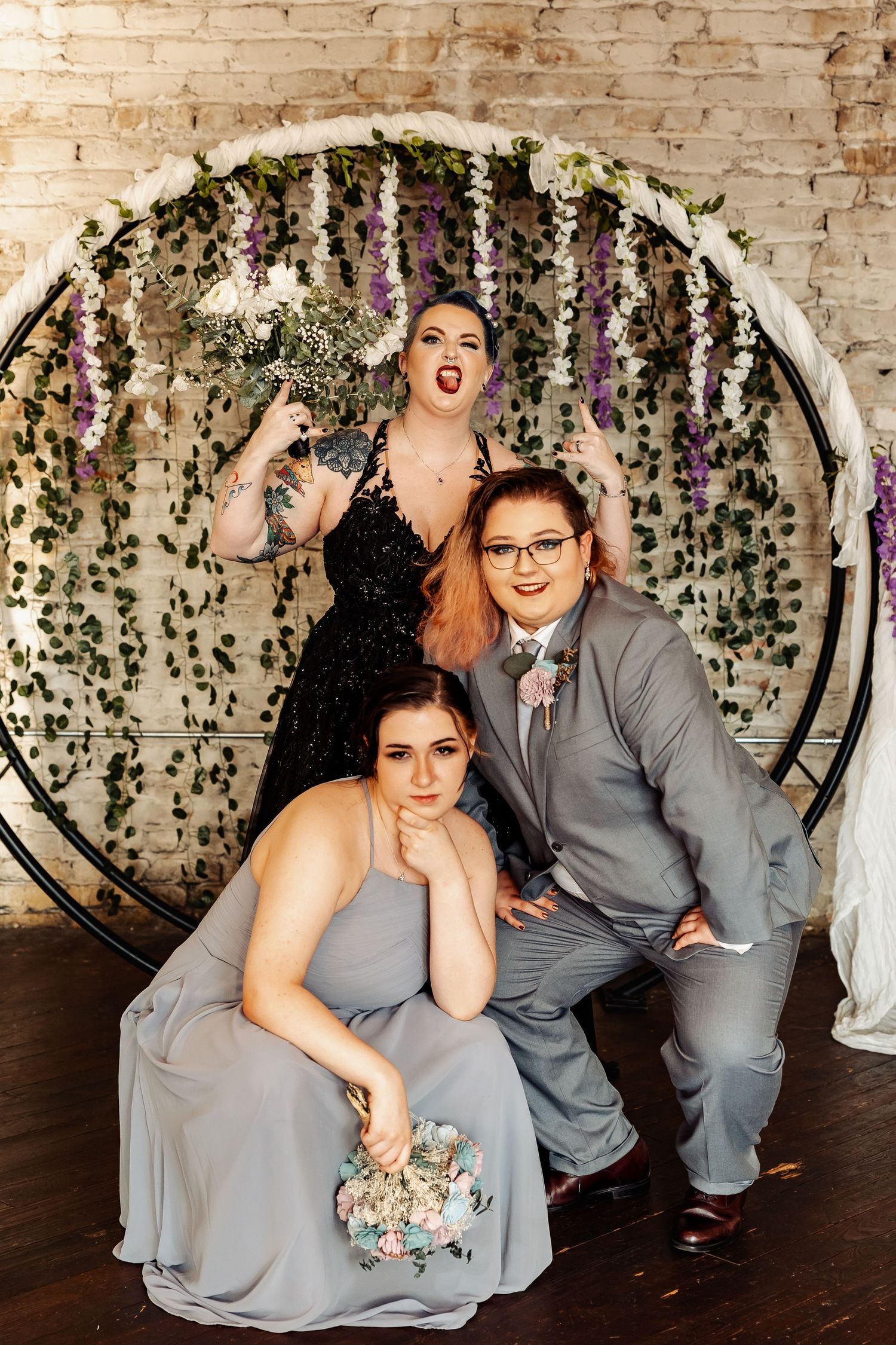 Three wedding party members pose playfully before a circular floral arch with wisteria and greenery.