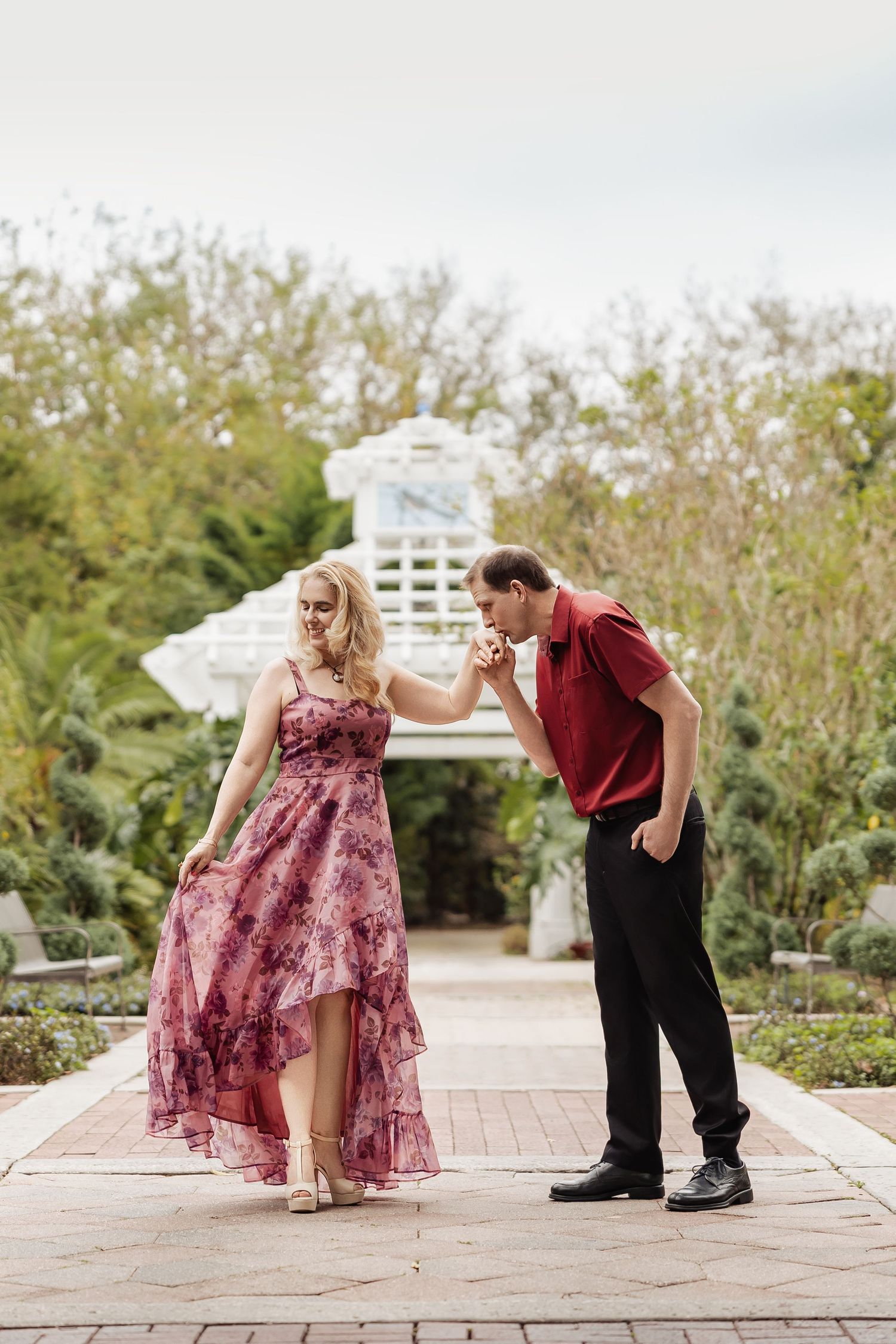 Man gallantly kisses womans hand as she wears a flowing floral pink dress in a garden setting.