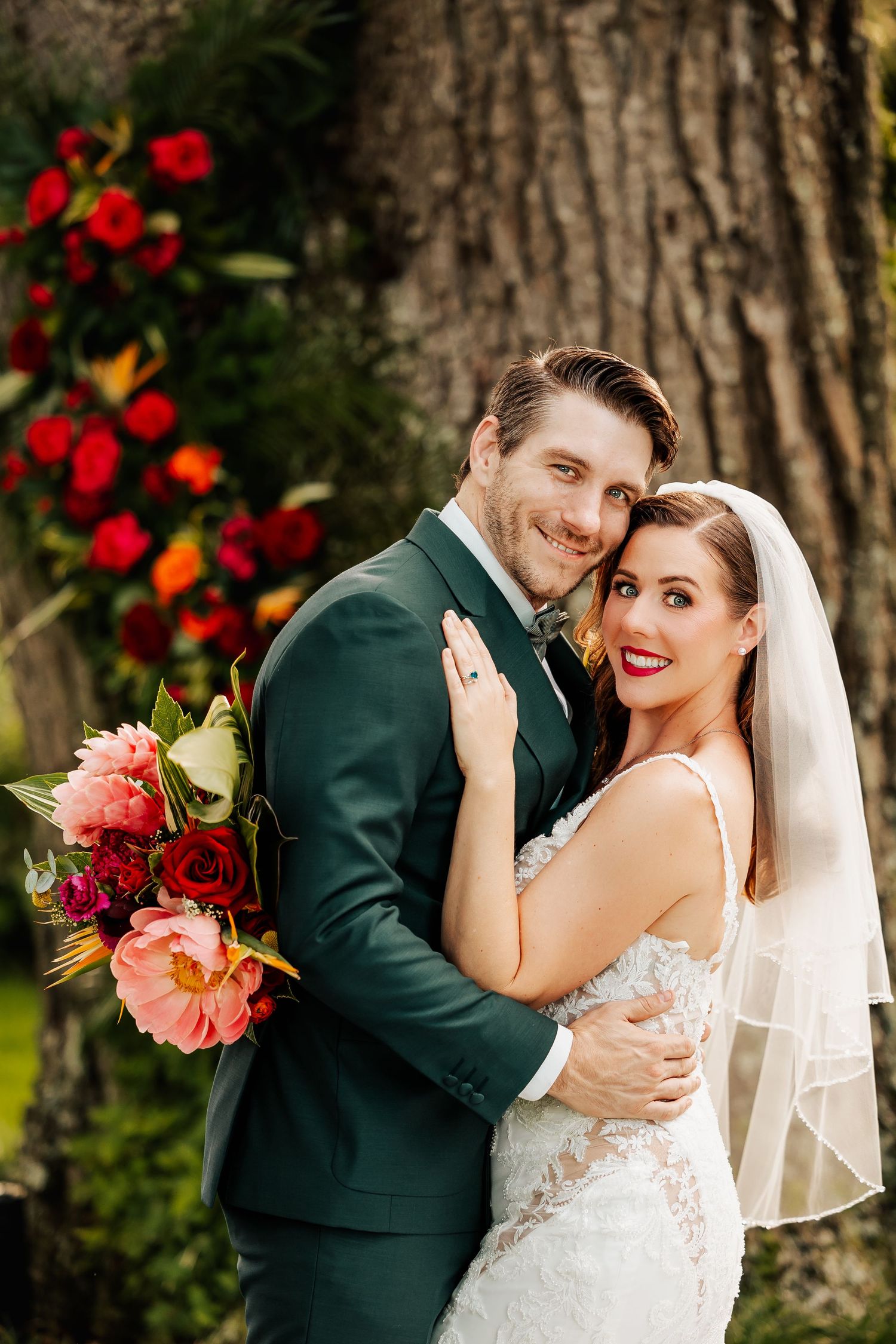 Bride and groom embracing before large tree with vibrant red floral arch at outdoor wedding.