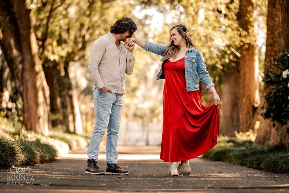 Couple sharing a playful moment on a tree-lined path, with woman wearing flowing red dress.