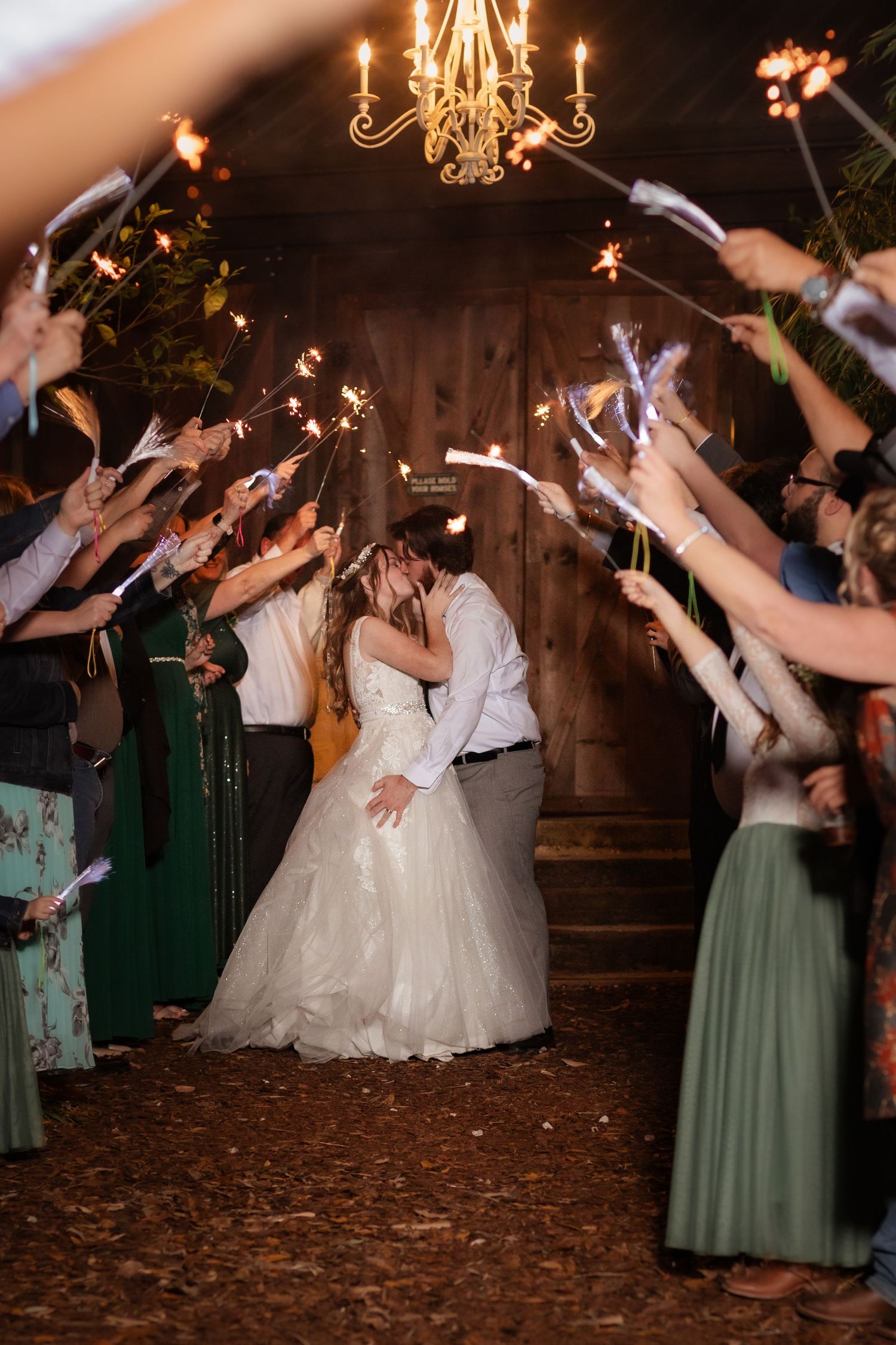 Wedding sparkler exit with couple kissing under chandelier surrounded by guests.