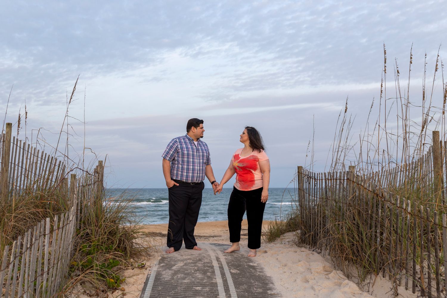 Couple holding hands while walking on beach boardwalk through sea oats at sunset.