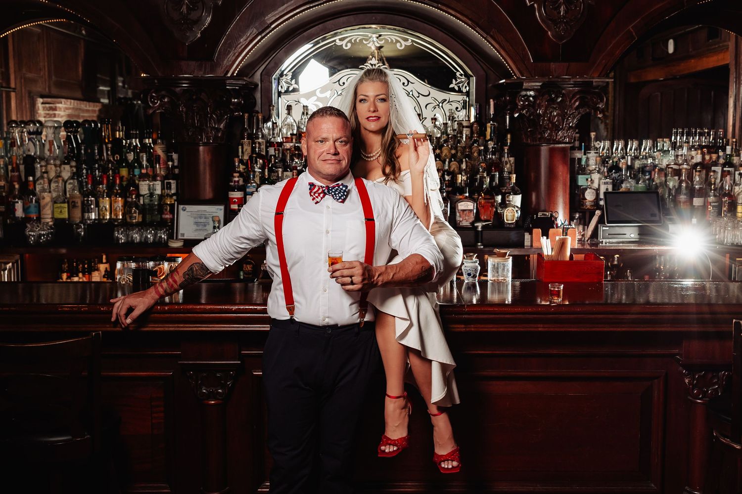 Bride and groom posing at a vintage wooden bar, groom in red suspenders and bow tie, bride in white dress.