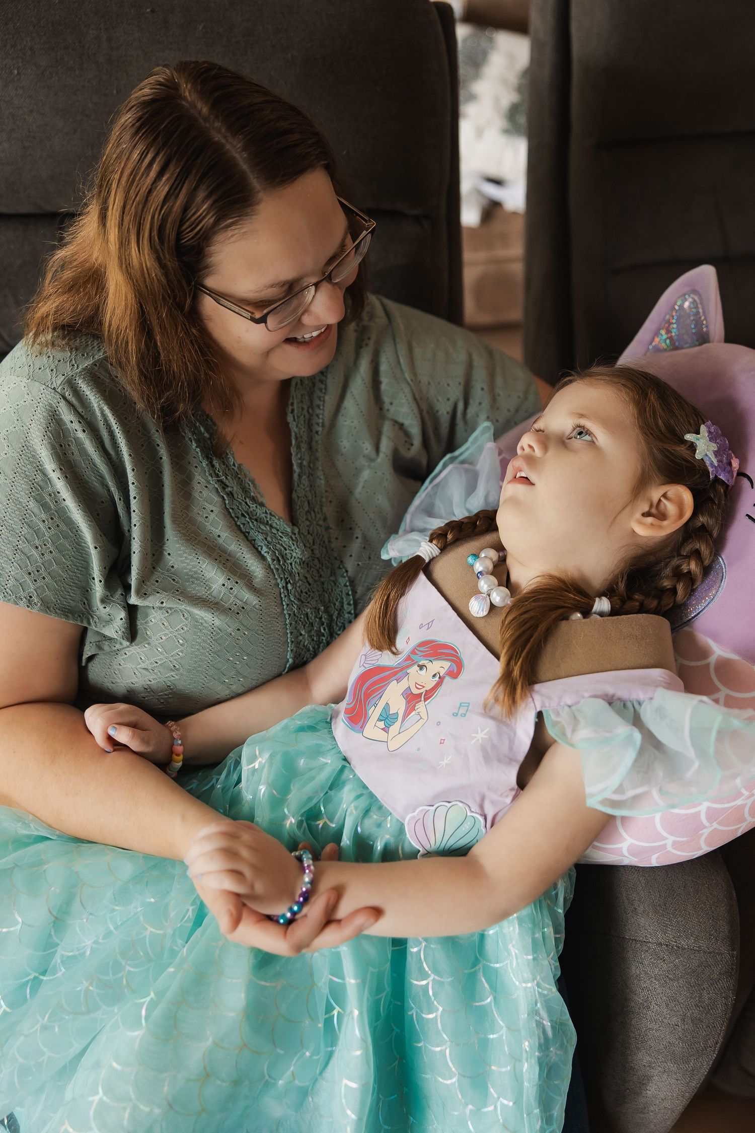 Mother smiling at her young daughter wearing a mermaid dress and fairy wings indoors.