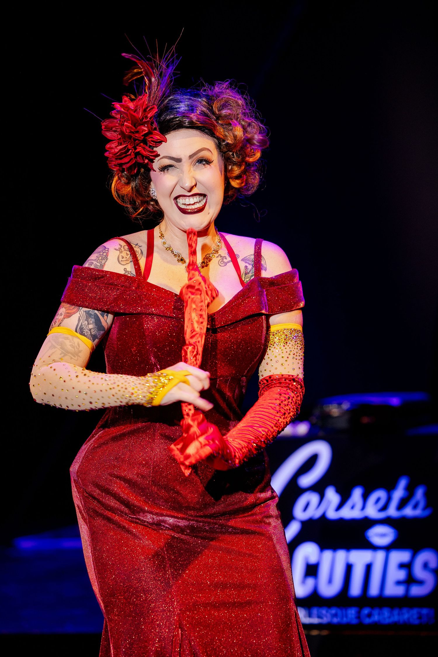 A smiling burlesque performer in a red sequin gown with feathered headpiece performs at Corsets and Cuties Cabaret.