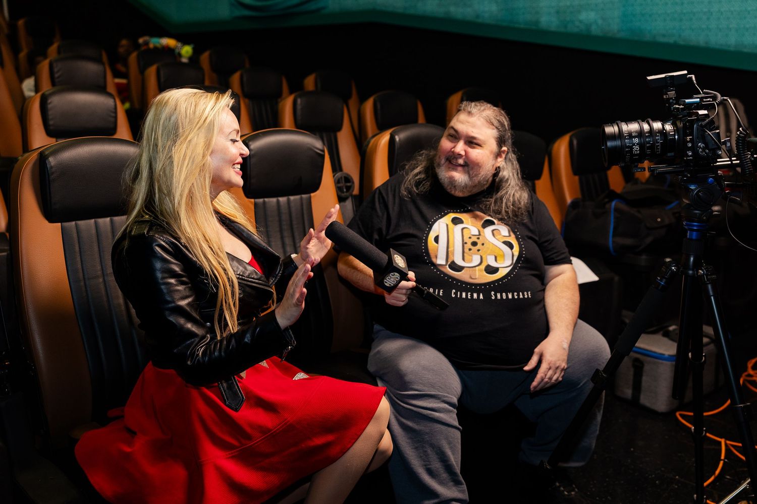 A blonde woman in a red skirt is interviewed on camera inside a luxury cinema screening room.