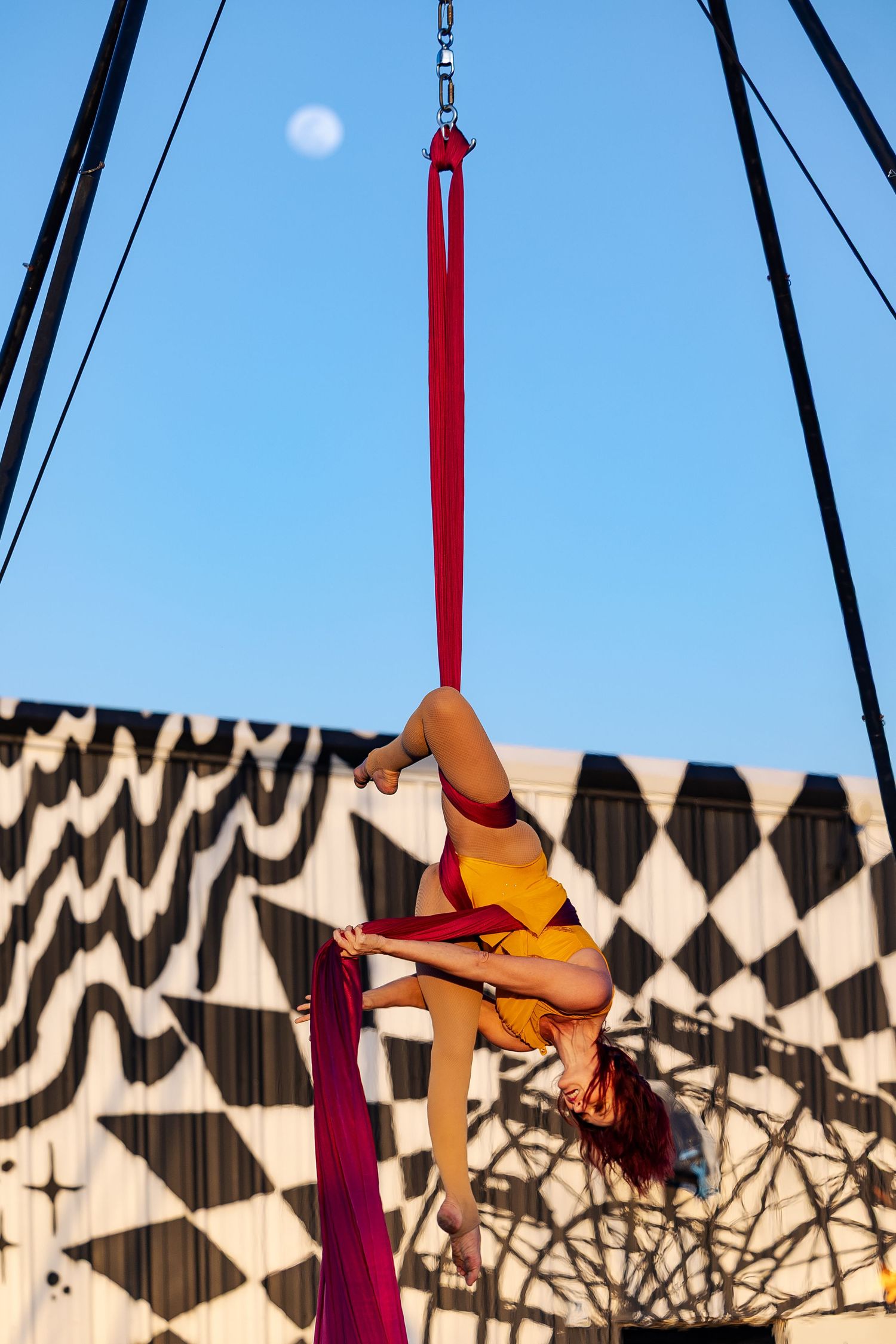 Female aerialist in yellow costume performing graceful moves on red aerial silks against a blue sky.