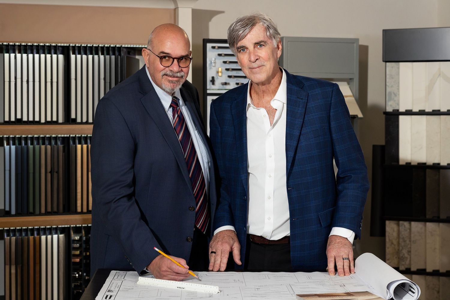 Two professional men reviewing architectural blueprints in a tile and material showroom.