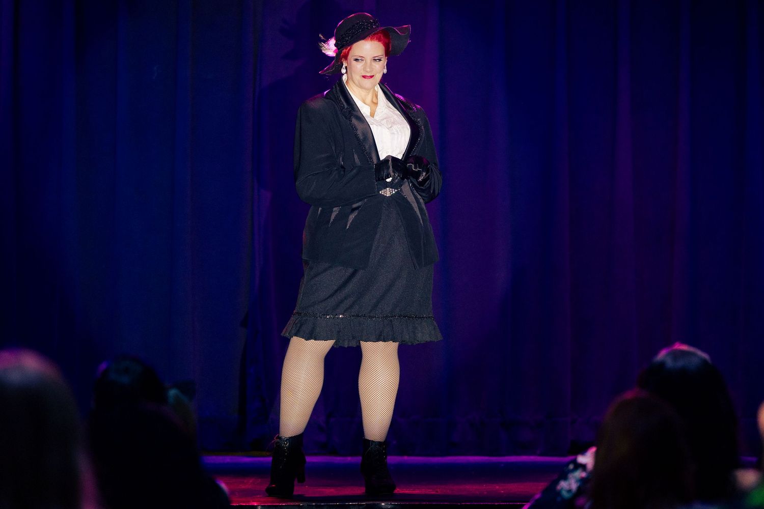 A woman in a black trench coat and boots performs on a lit stage during a burlesque show.