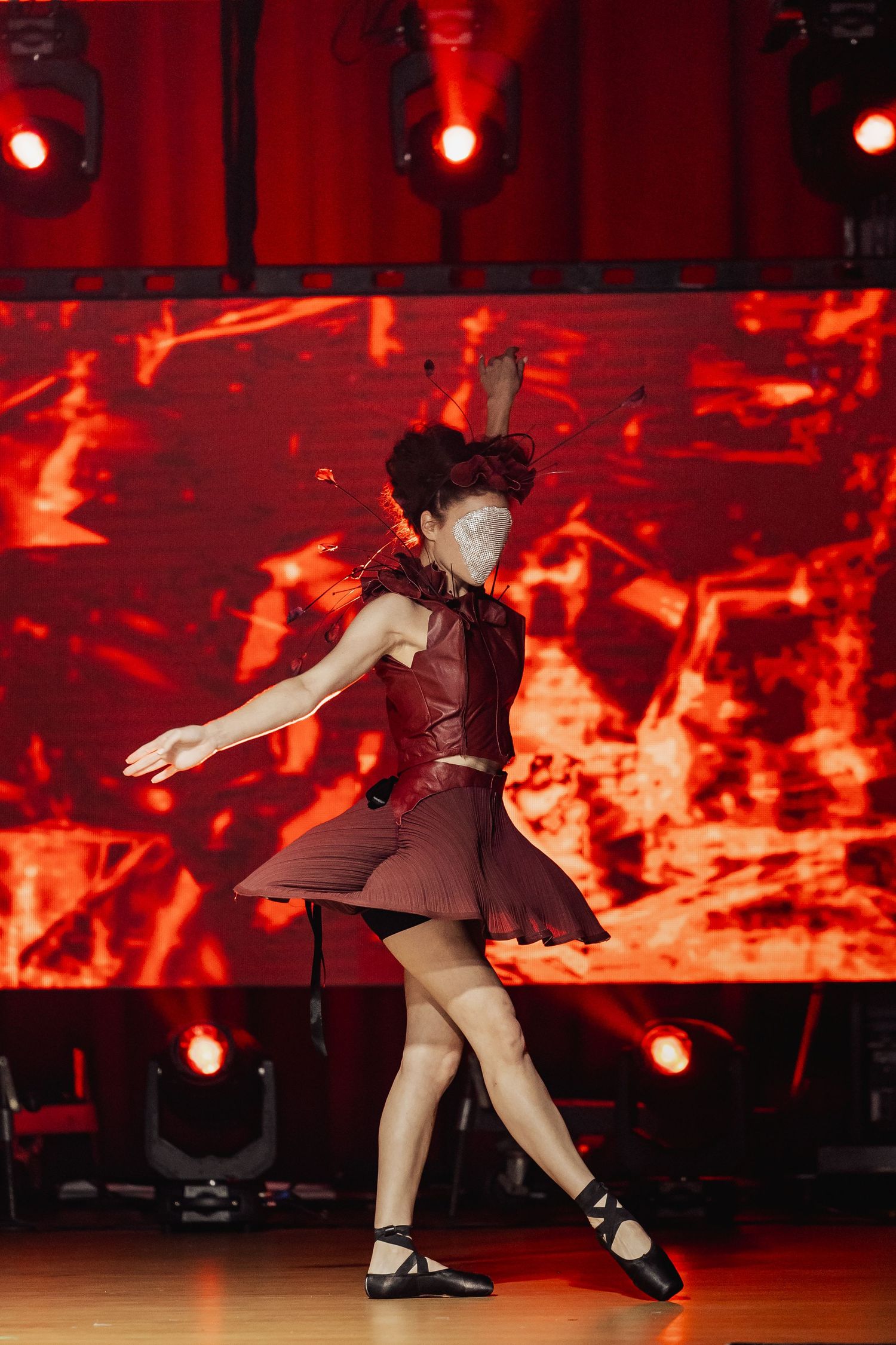 Dancer in dark red leather outfit with decorative headpiece performs on stage with dramatic red backdrop.
