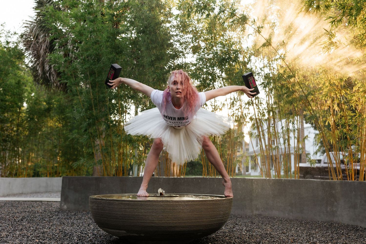 Ballerina in white tutu performing a handstand on a decorative bowl fountain surrounded by bamboo.