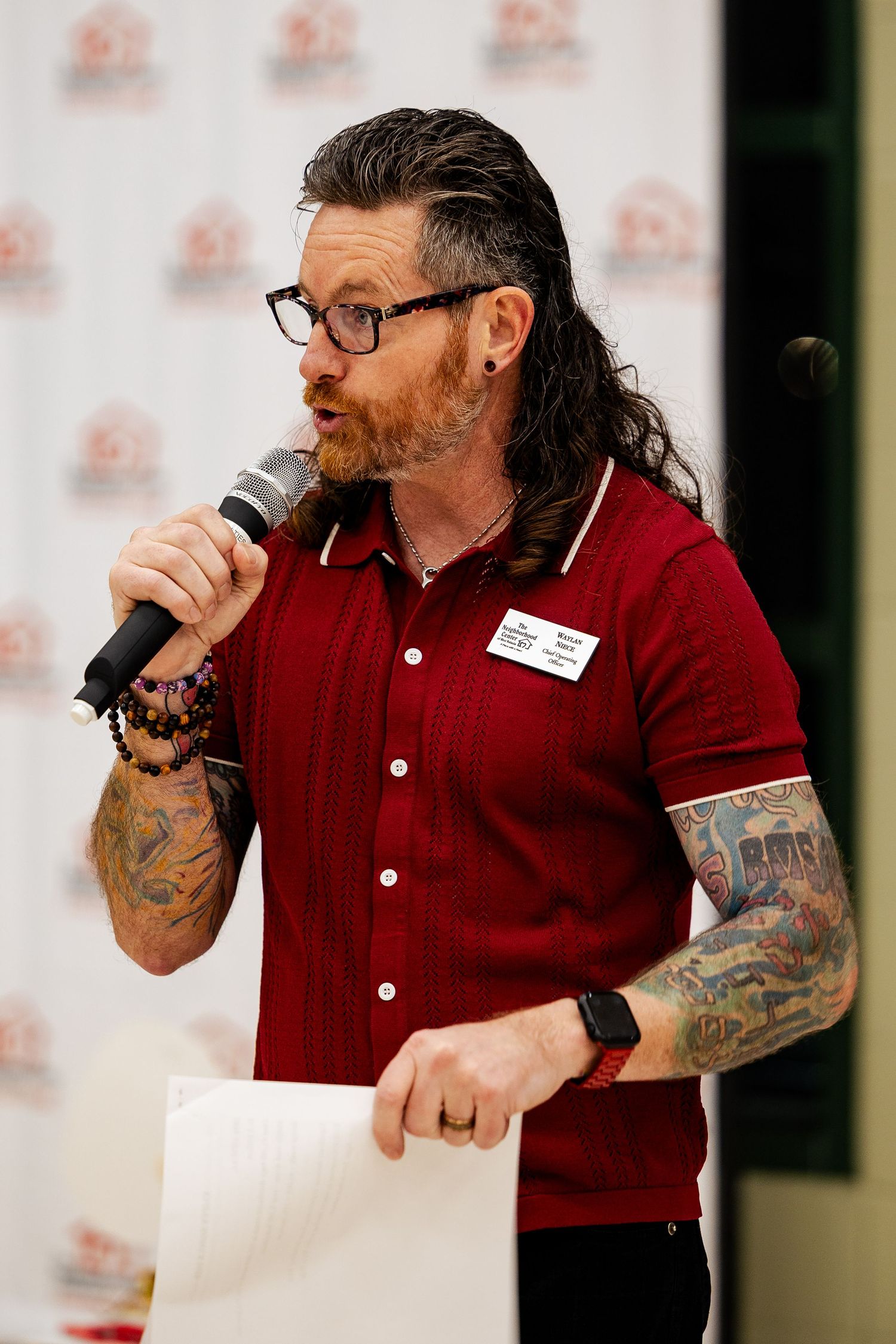 A tattooed man in a red shirt speaks into a microphone at a community fundraising event.